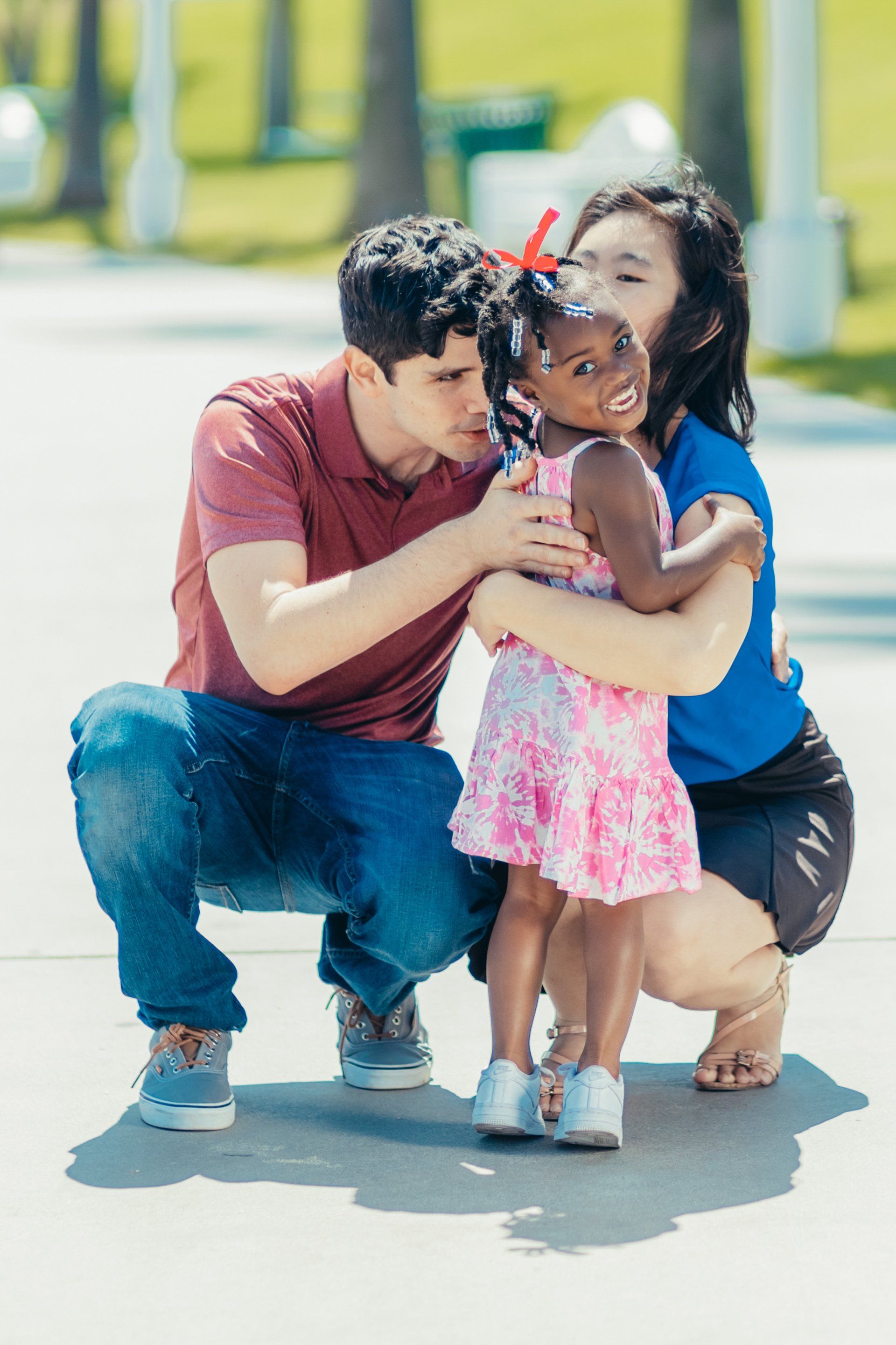 A man and a woman in casual clothing crouch to hug a young child in a pink patterned dress on a paved outdoor path.
