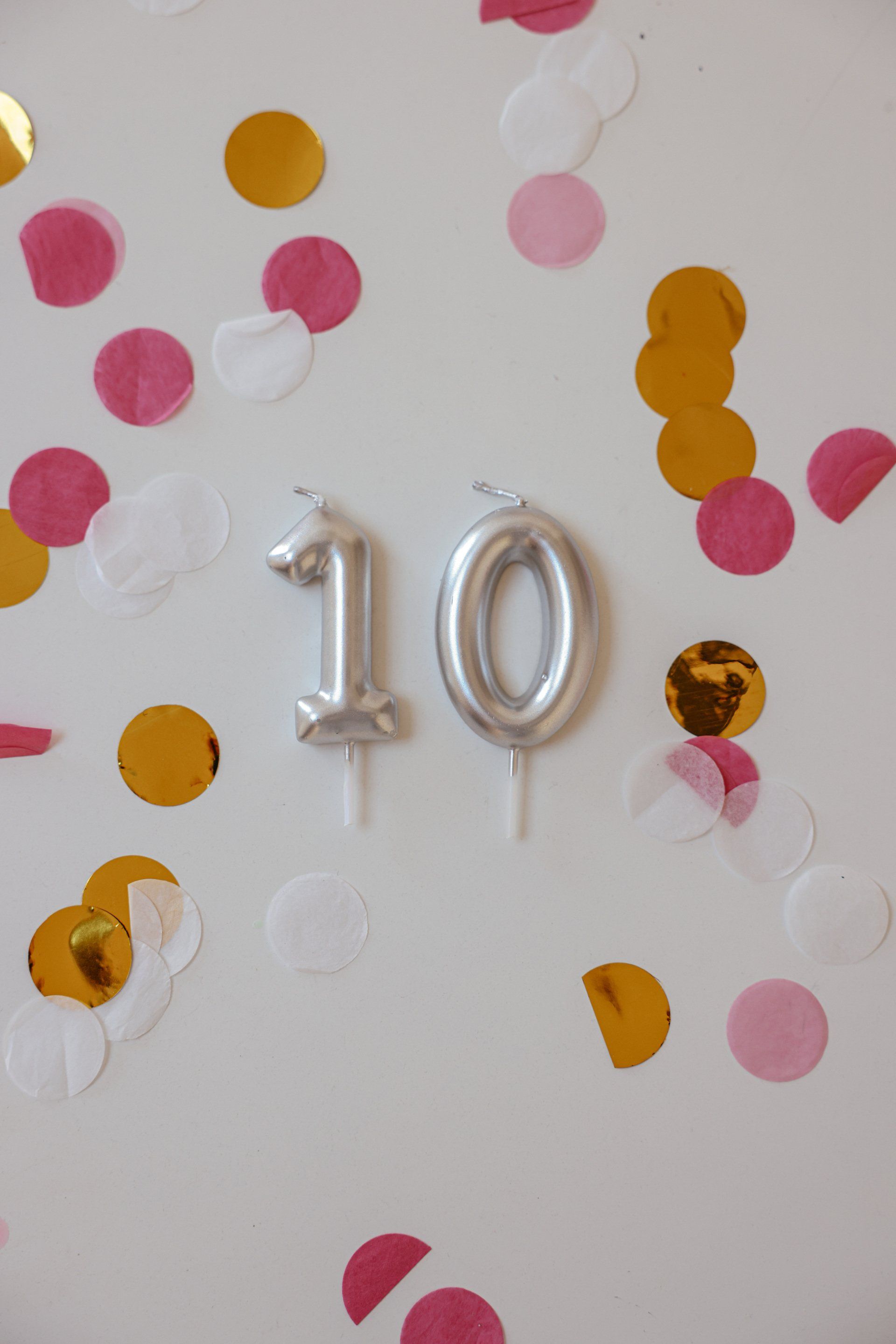 Silberne Geburtstagskerzen mit der Zahl „10“, umgeben von goldenem, rosa und weißem Konfetti an einer weißen Wand.