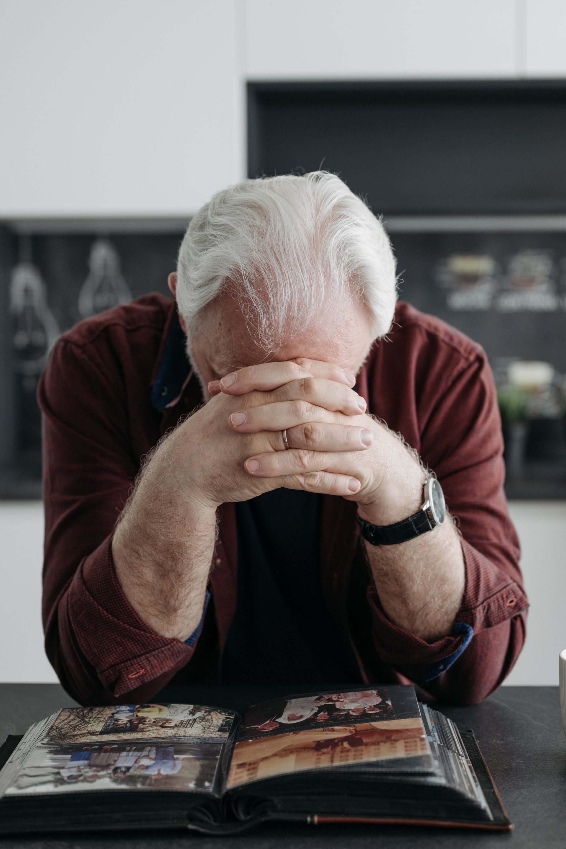 An older man is sitting at a table with his head in his hands and looking at a photo album.