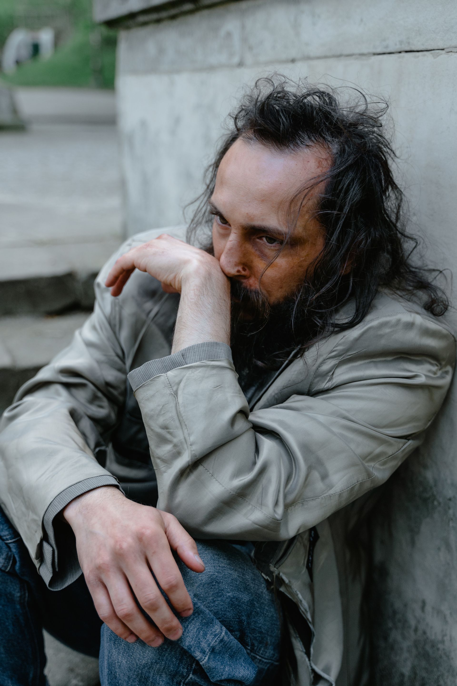 Man sitting, leaning against a concrete structure, looking down with a sad expression.