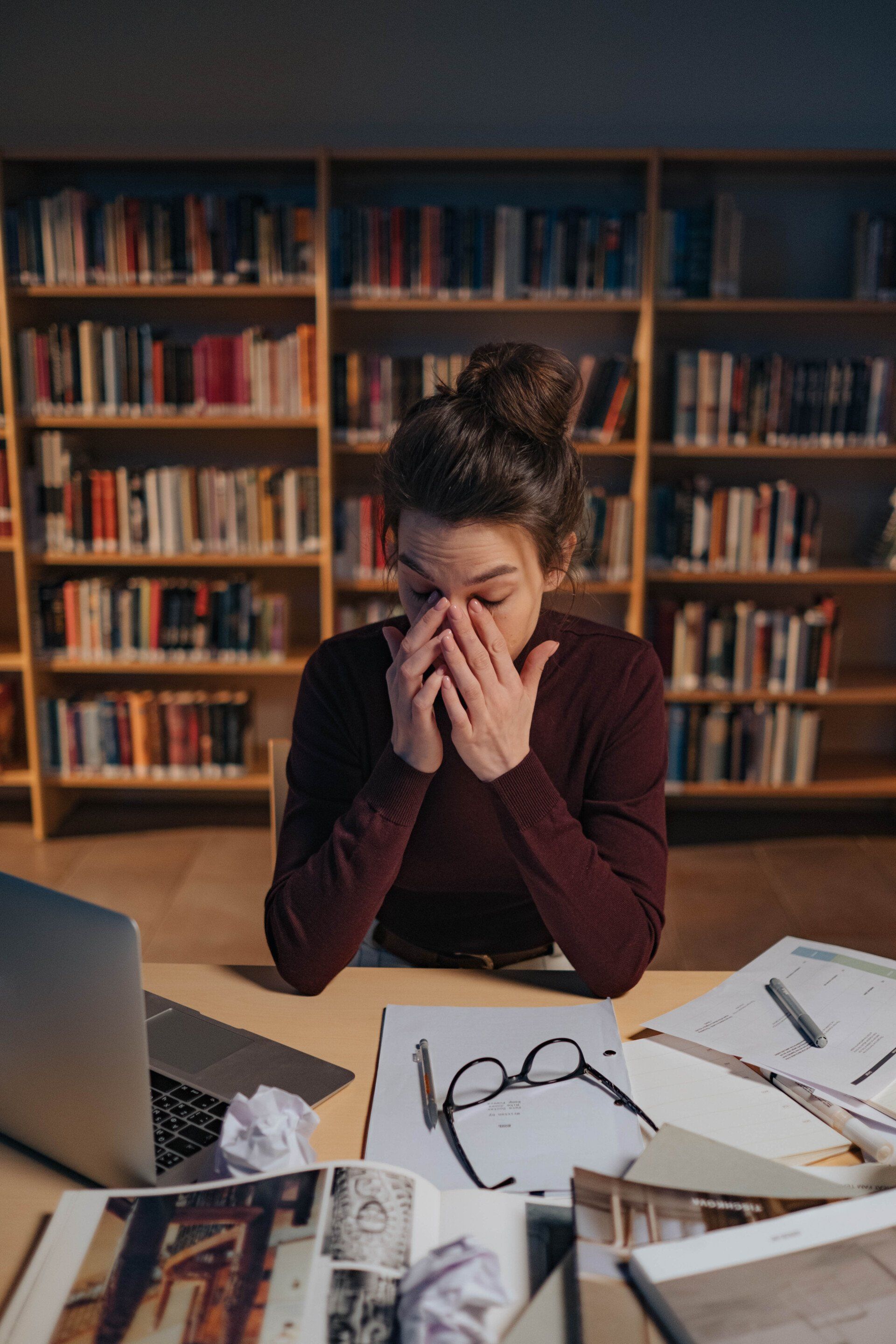 A woman is sitting at a desk in a library covering her face with her hands.