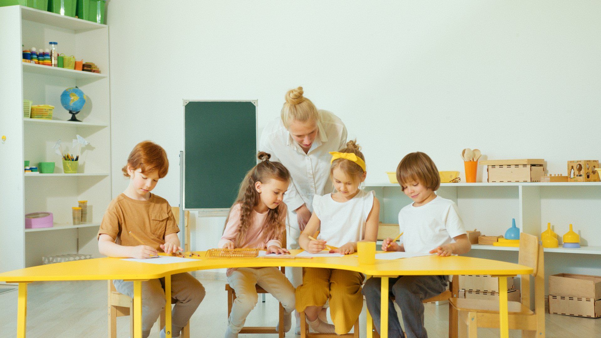 Kinderen tekenen in een klaslokaal aan een gele tafel; de leerkracht helpt.