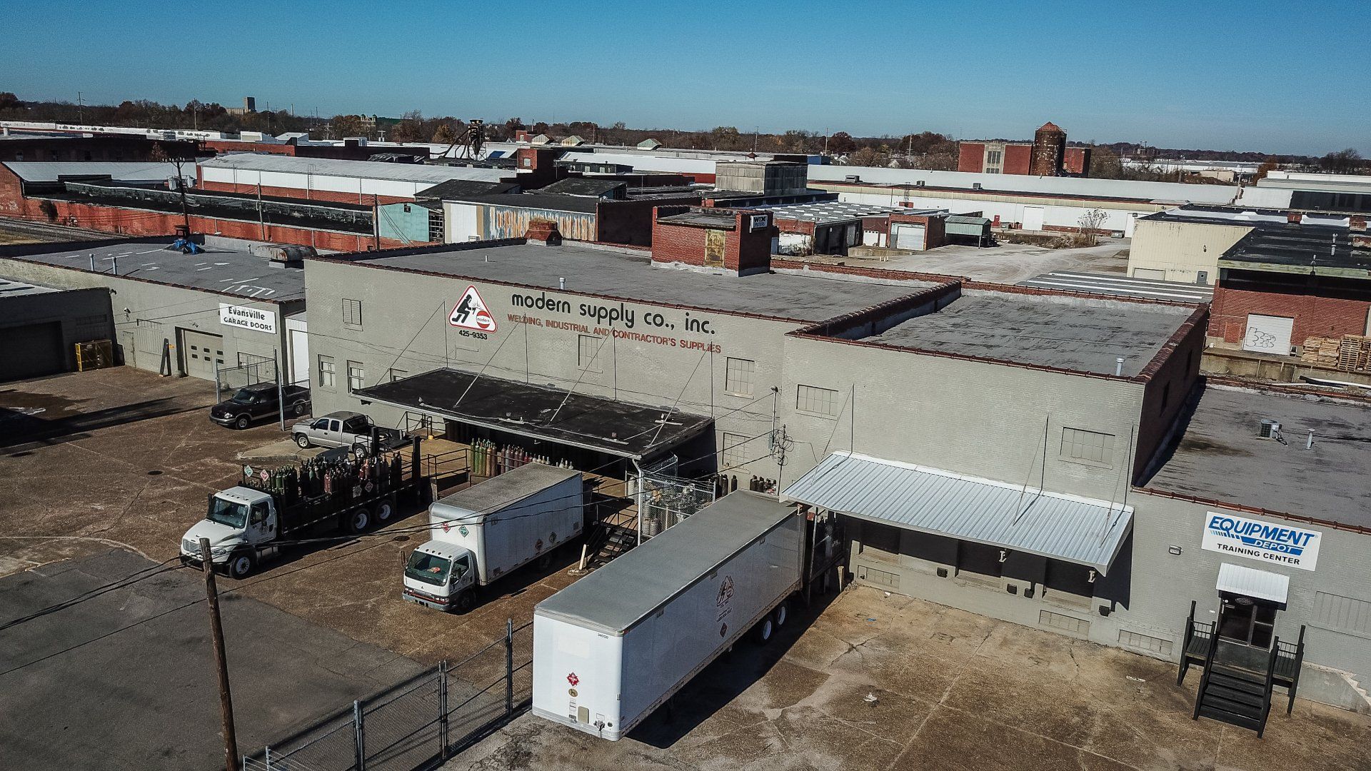 An aerial view of a gray industrial building with a flat roof, loading docks, white trucks, and surrounding urban debris.
