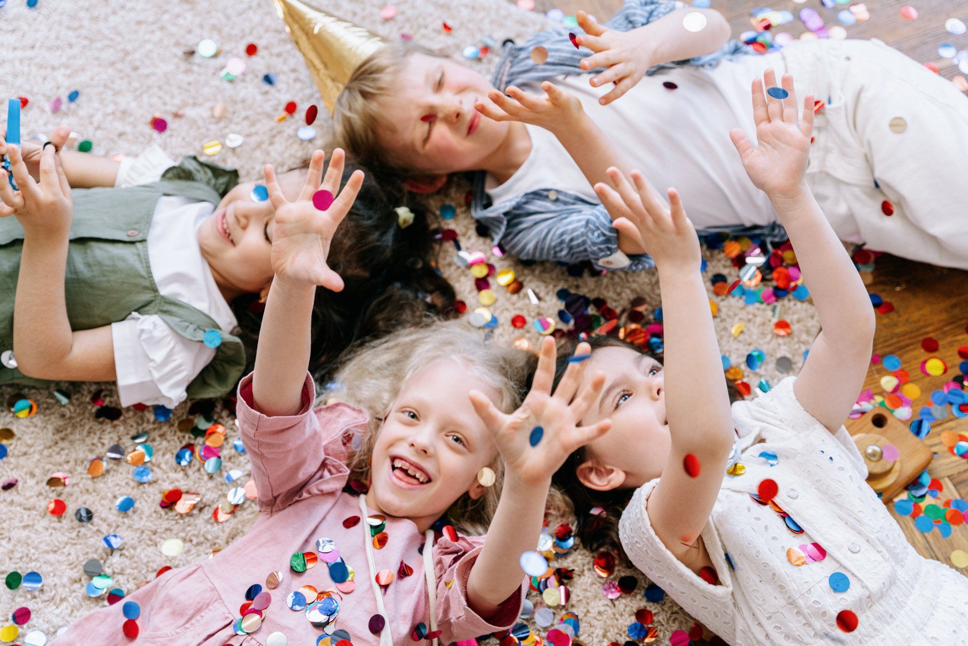 Children lie on a floor covered in confetti, smiling and raising their hands.