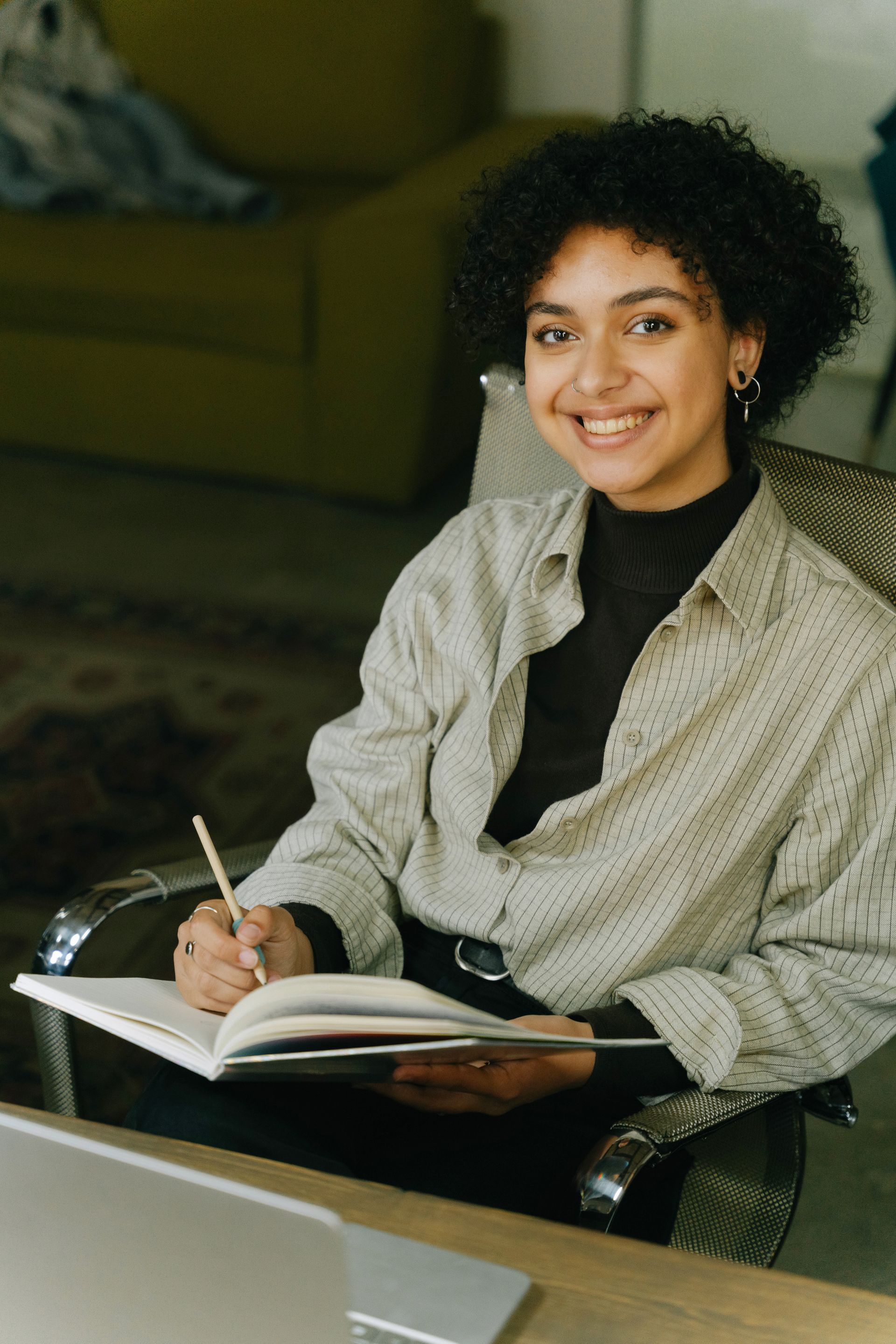 A woman is sitting at a desk writing in a notebook and smiling.