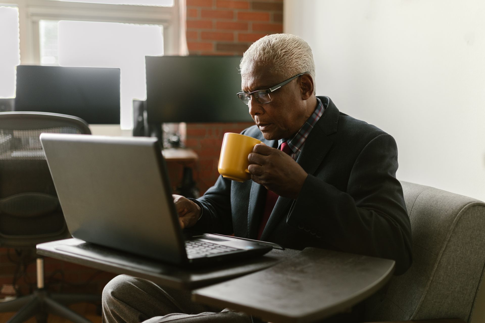 Man in blazer sips from mug, looks at laptop, and sits in an office setting.