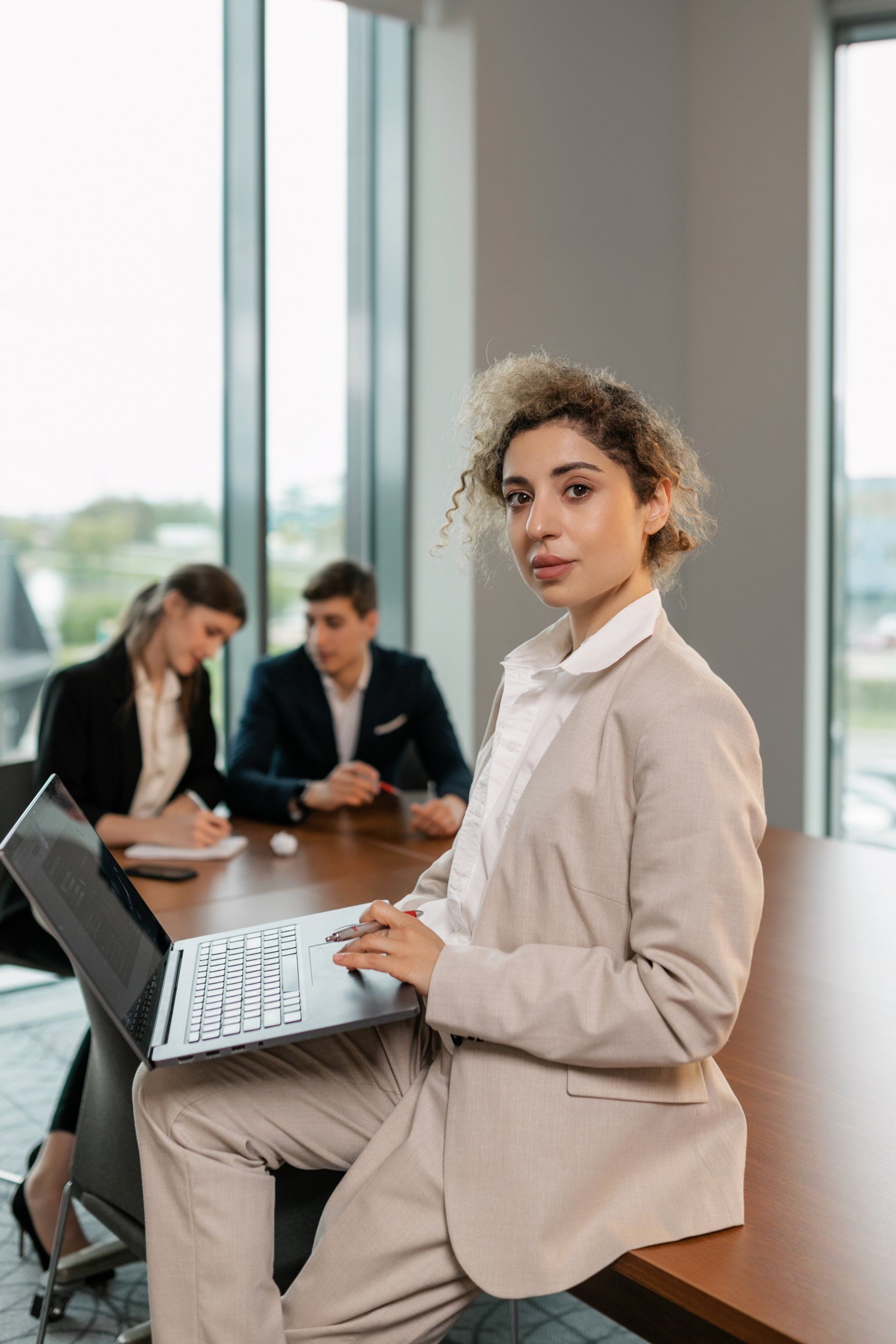 Woman in beige suit, perched on table, looks at camera, laptop in lap. Two colleagues in background.