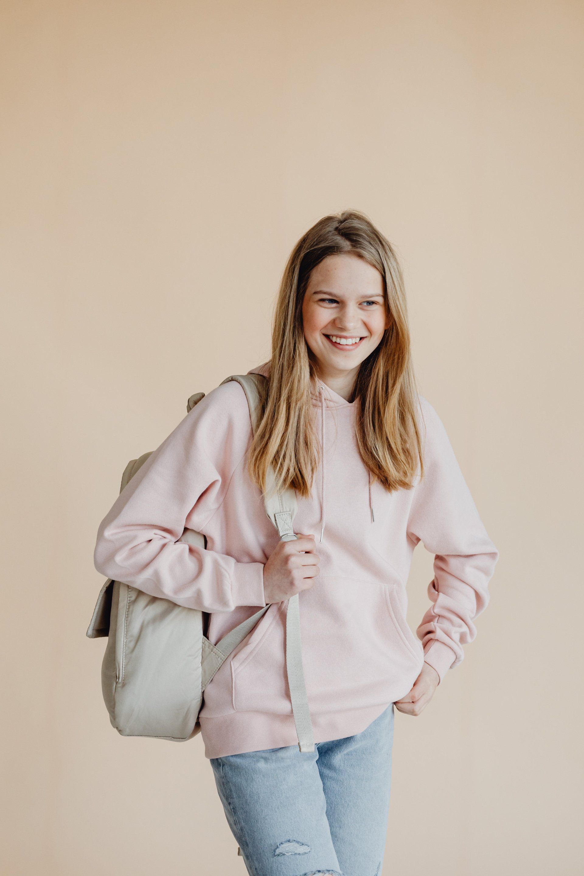 Persoon in roze hoodie en spijkerbroek met rugzak, lachend, tegen een beige achtergrond.