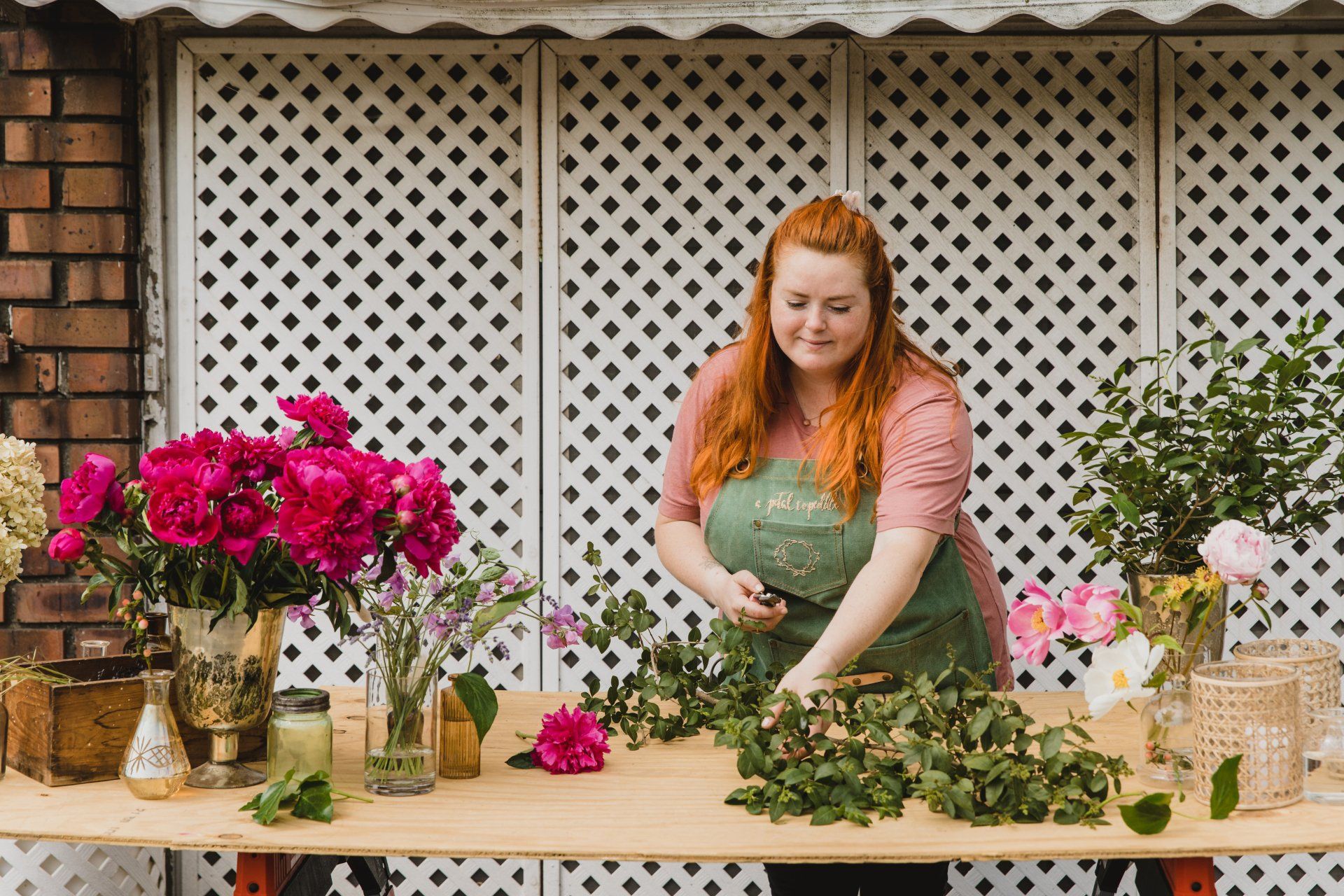 Woman arranging flowers at a wooden table; pink peonies, brick wall background.