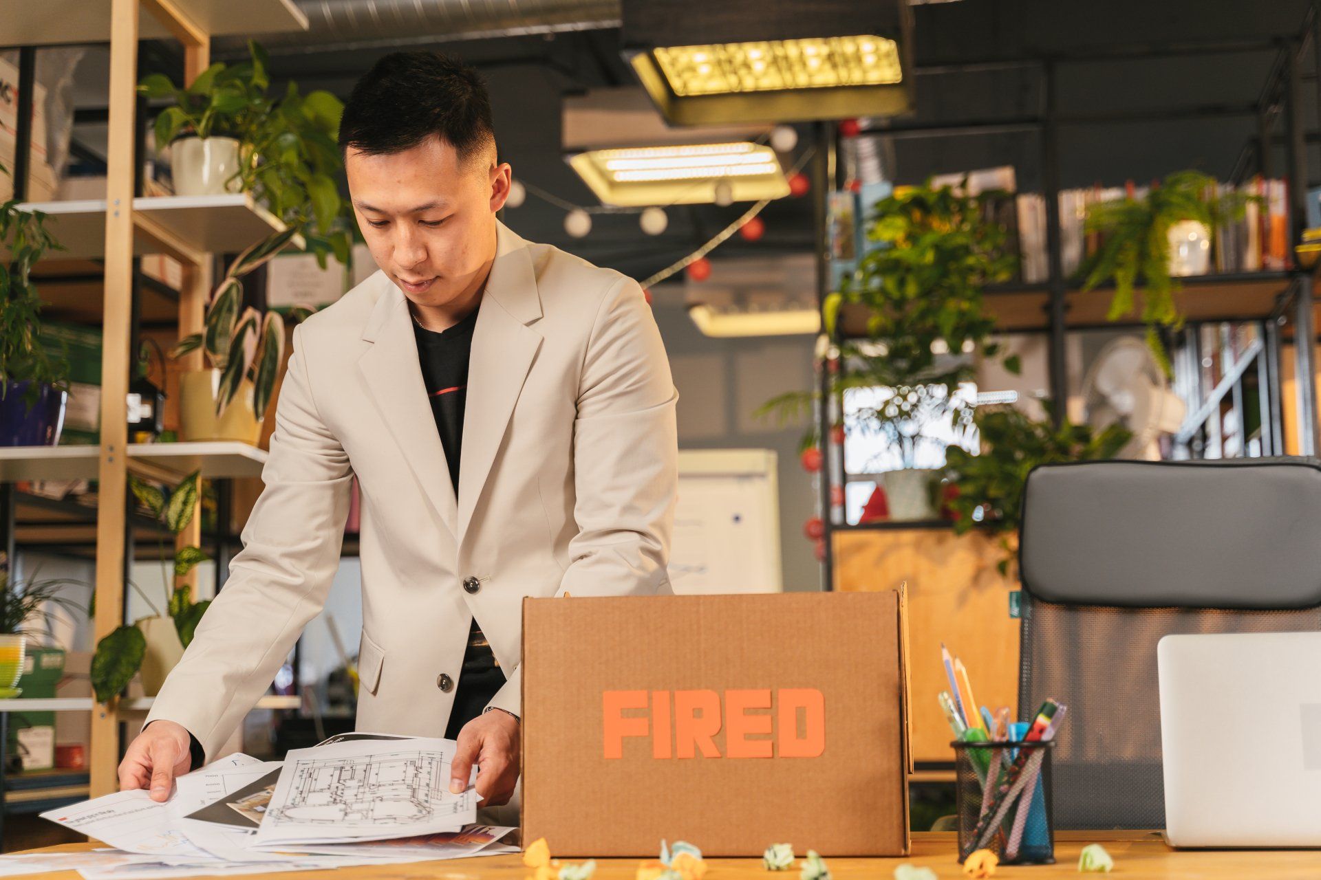 A person in a beige blazer packs documents into a cardboard box labeled "FIRED" on an office desk filled with plants.
