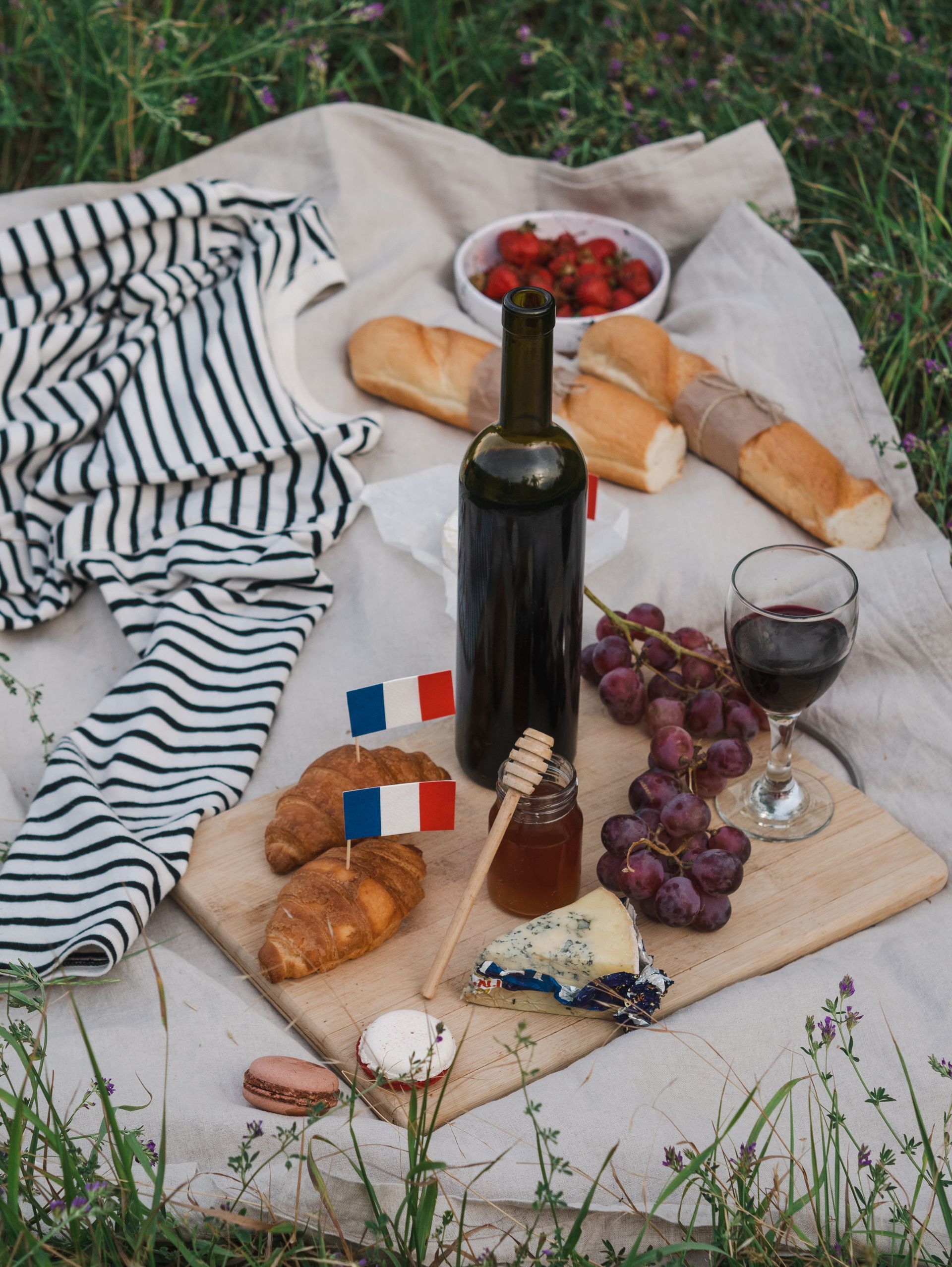 A picnic blanket with a bottle of wine , grapes , cheese , croissants and strawberries on it.
