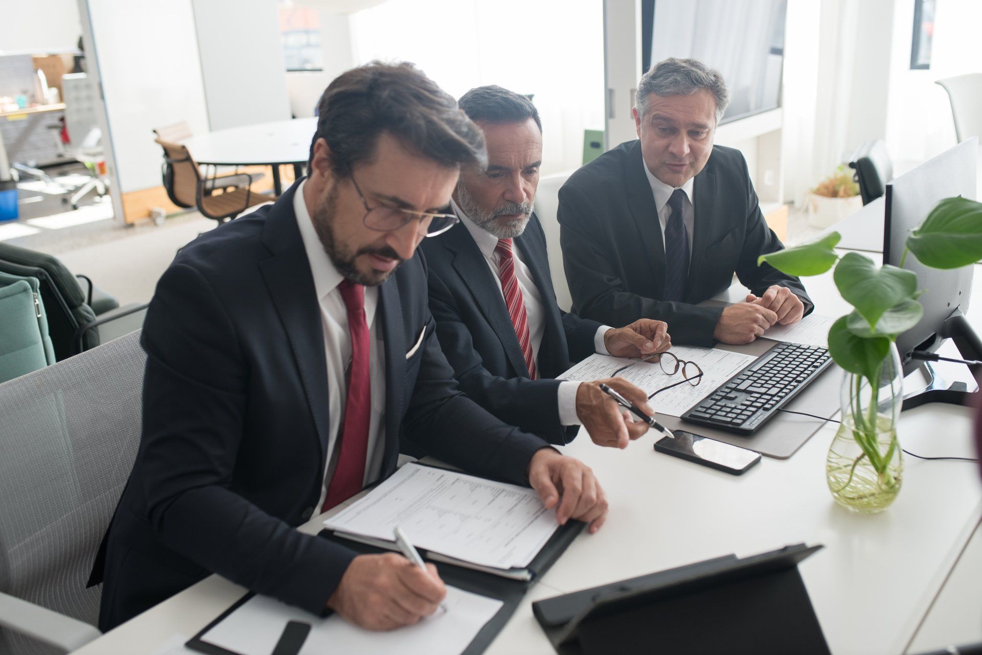 Three men in suits reviewing documents at a table in an office.