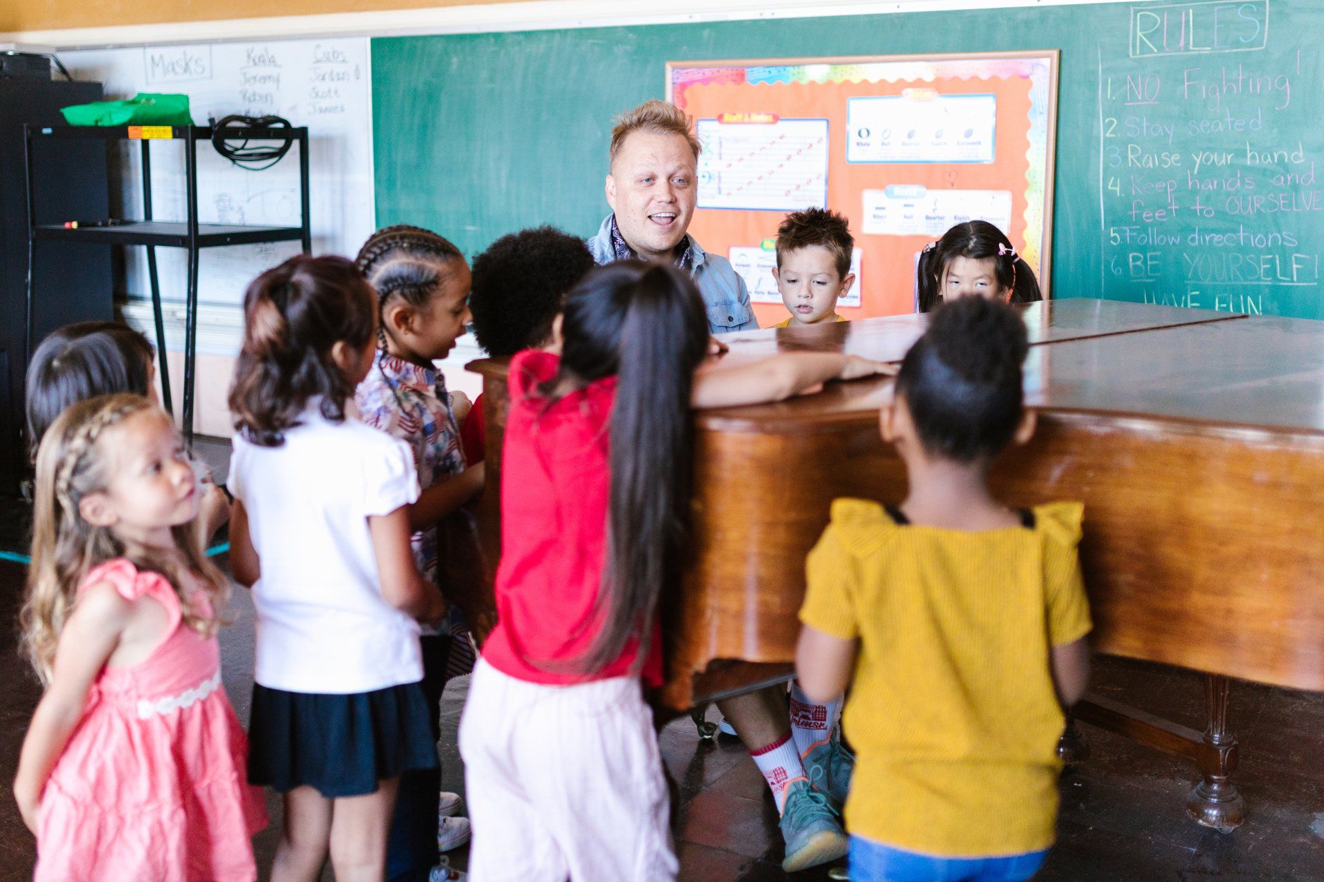 Students gathered around a teacher in a classroom, listening at a wooden desk with a chalkboard behind them