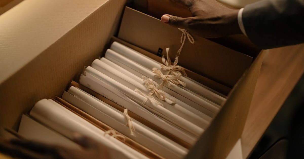 A person opening a cardboard box filled with bundled, rolled documents tied with twine.