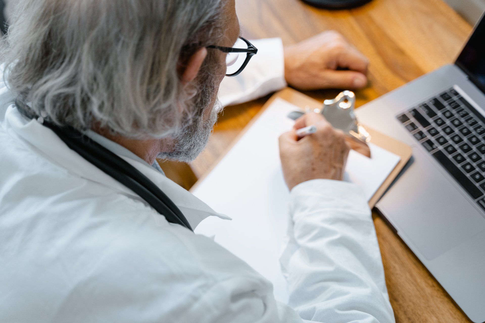A doctor is sitting at a desk with a clipboard and a laptop.