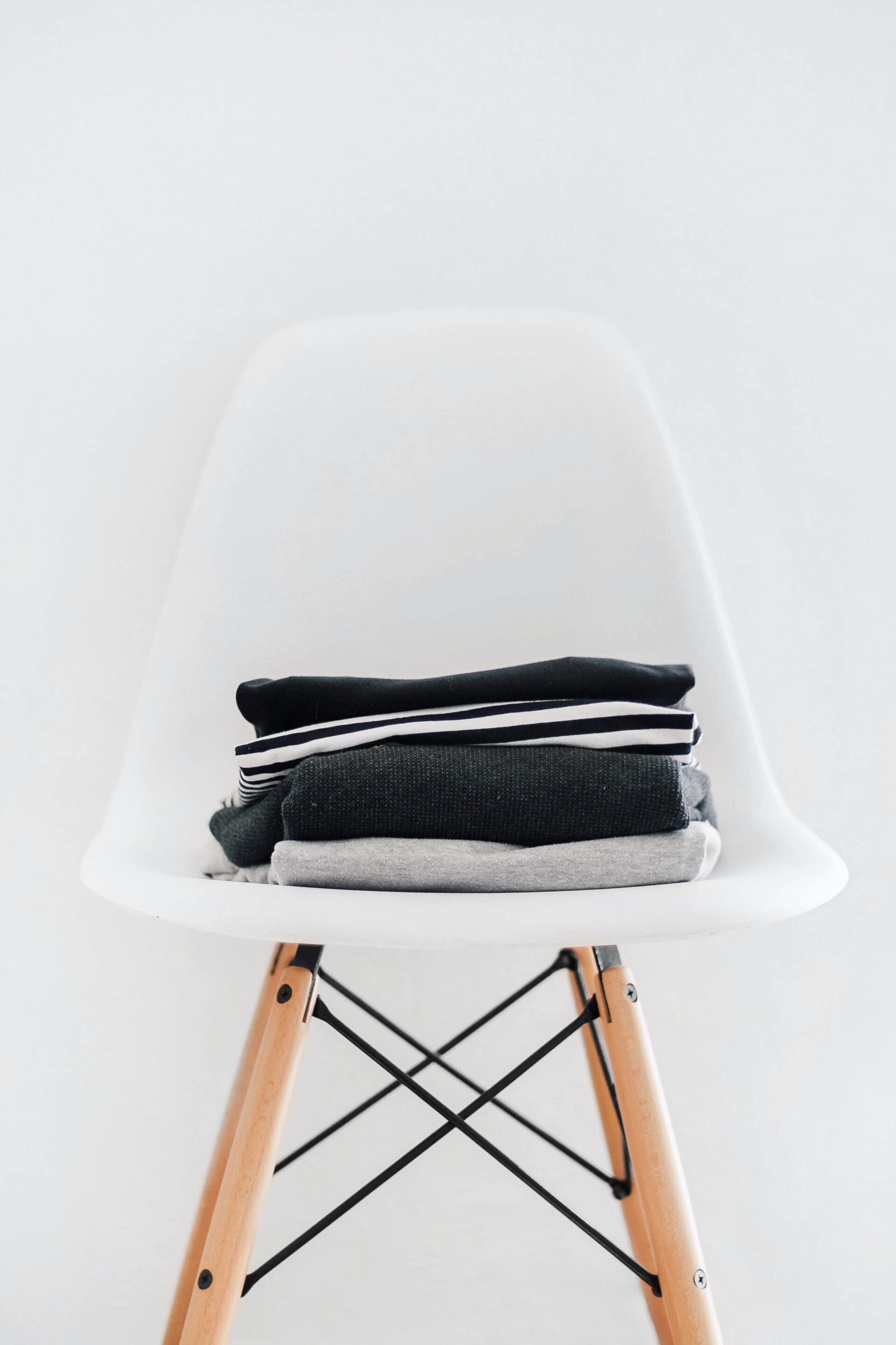 Stack of folded dark-colored clothes on a white chair with wooden legs, against a white wall.