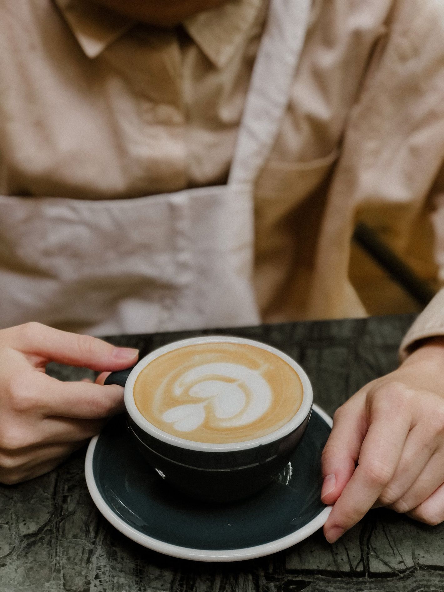 Hands holding a black cup of latte on a saucer, with beige clothing in the background