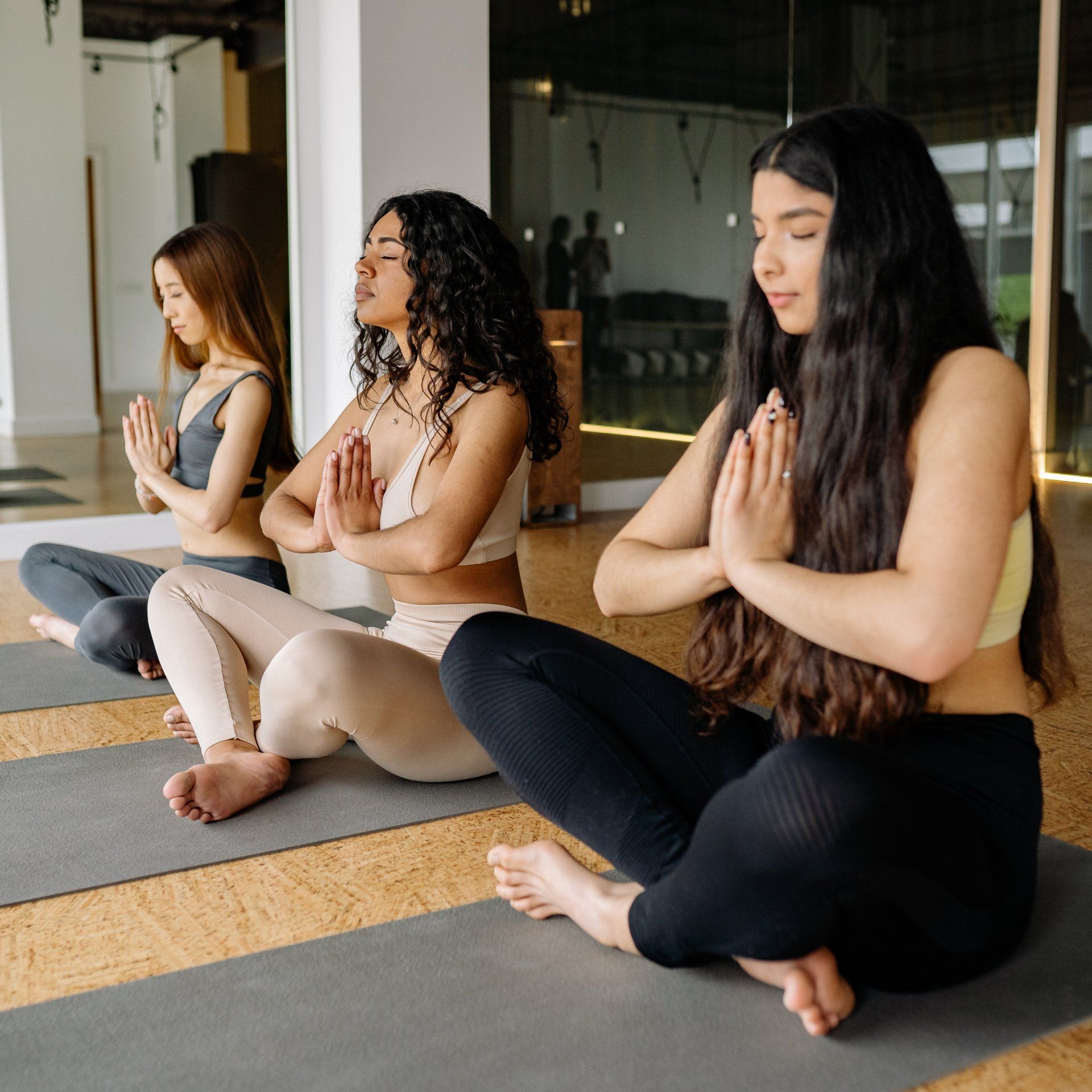 Three women in athletic wear seated in a yoga studio, hands in prayer, eyes closed.
