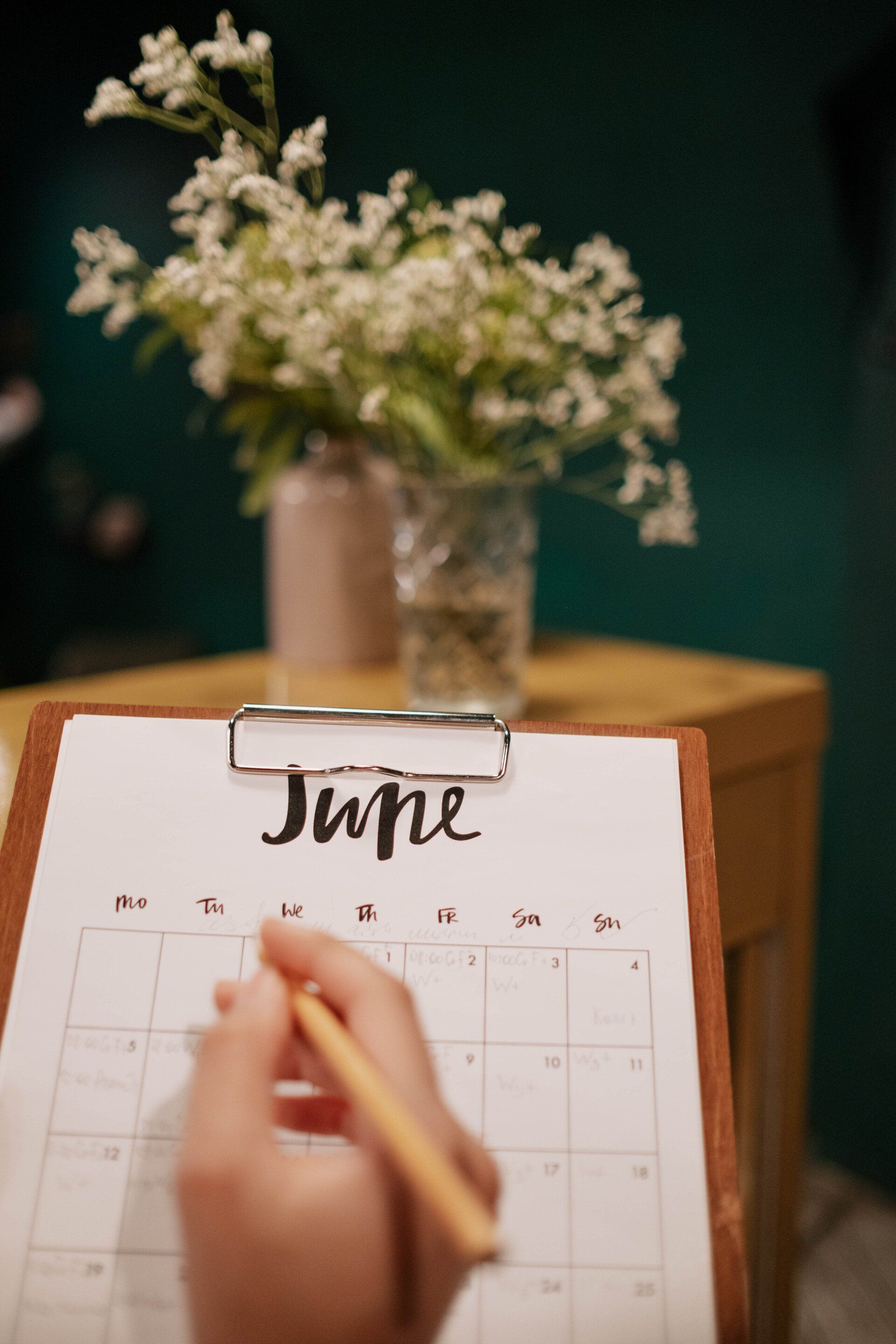 a person is writing on a clipboard with a pencil in front of a vase of flowers .