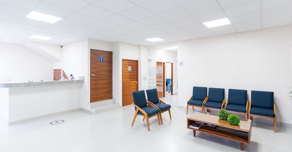 Waiting room with blue chairs, coffee table, and wood doors. Reception desk on the left. White walls and ceiling.