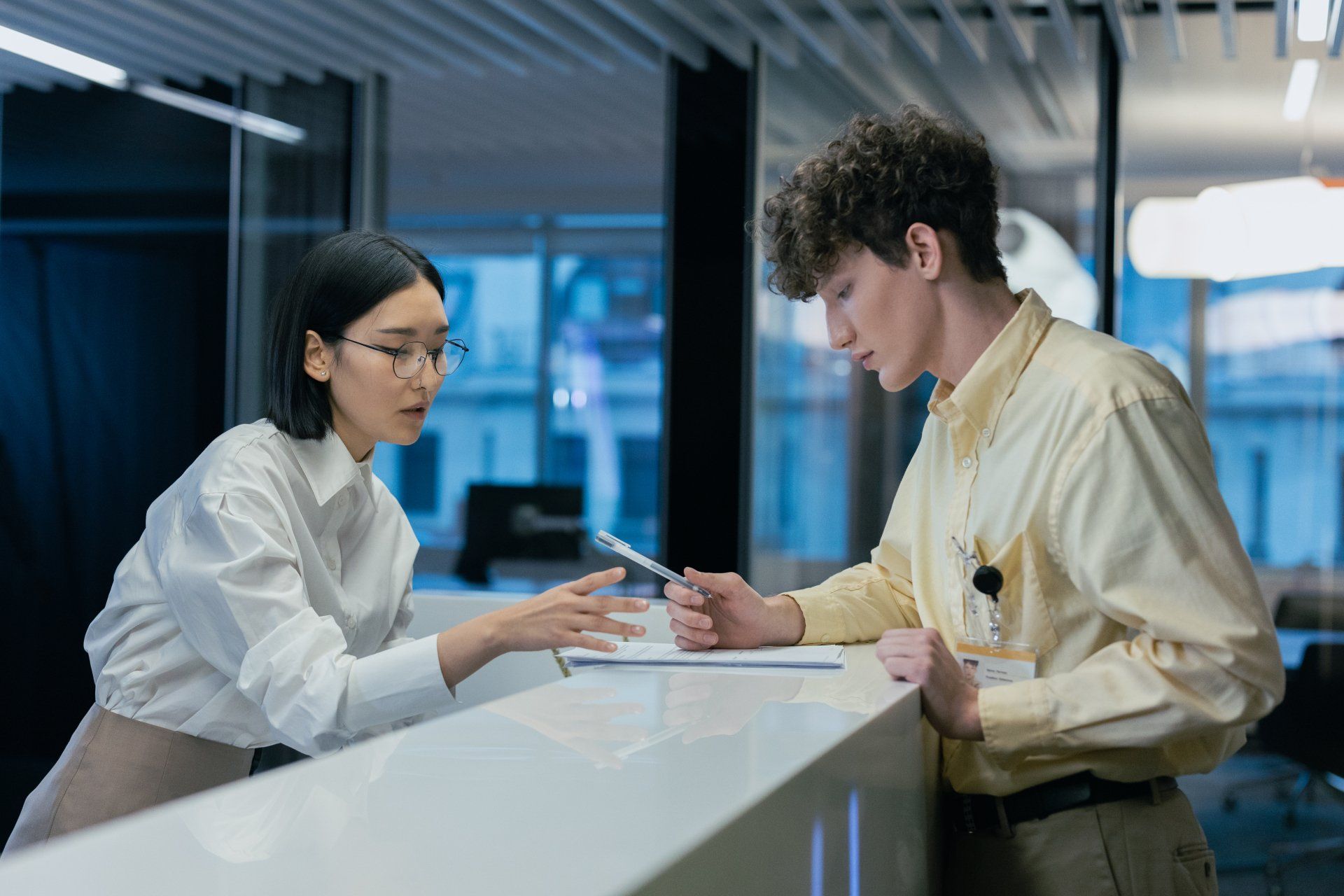 Woman with glasses points to document, man examines it at a front desk in an office.