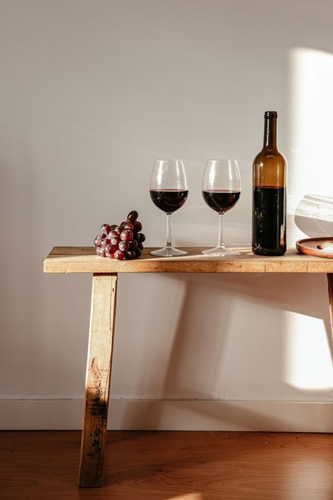 Two wine glasses and bottle with grapes on rustic wooden bench.