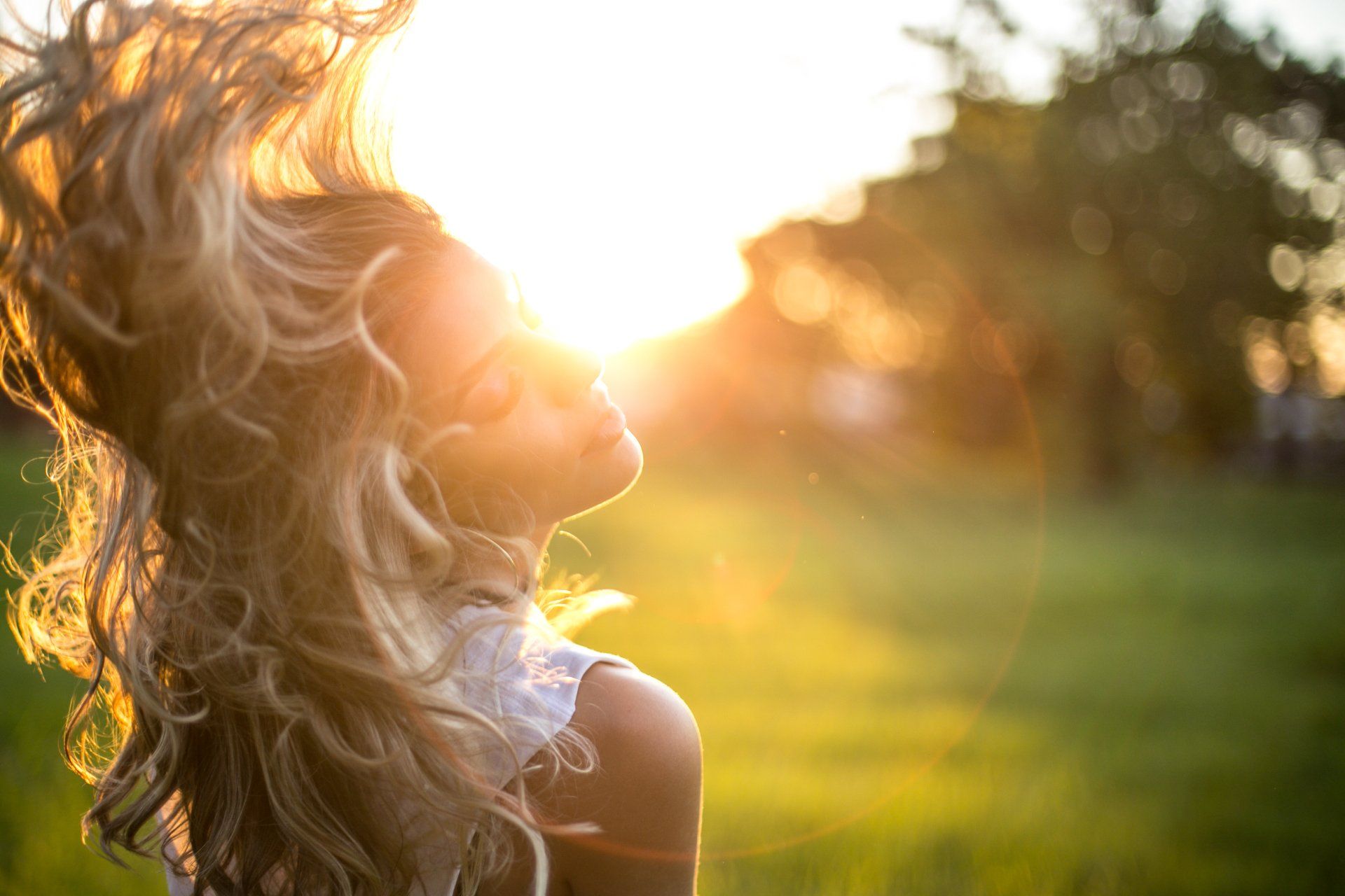 Woman with blonde hair backlit by the sun, looking upward with eyes closed, in a field.