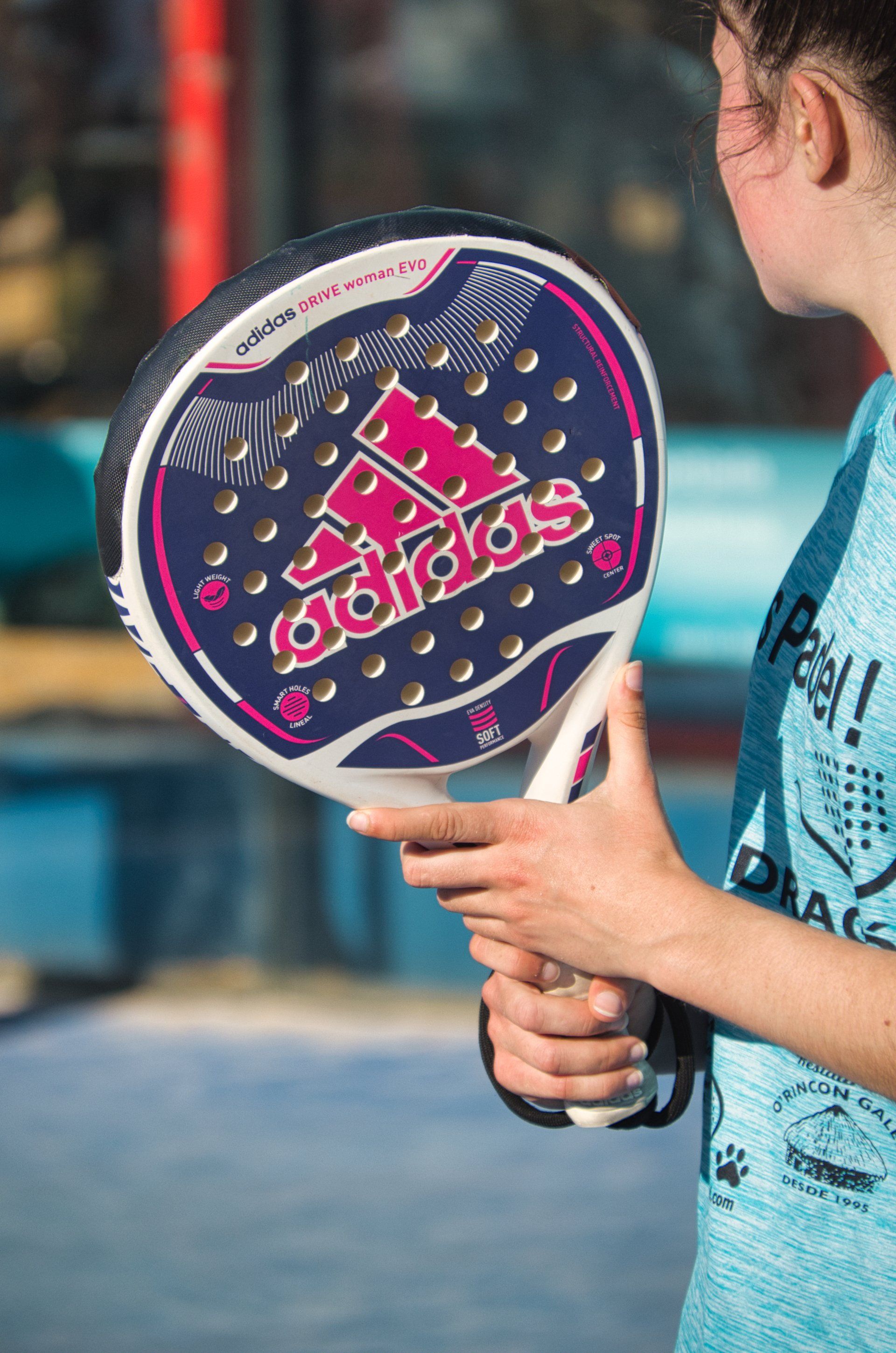 Een persoon met een Adidas padelracket met een donkerblauw, wit en roze design staat op een padelbaan.