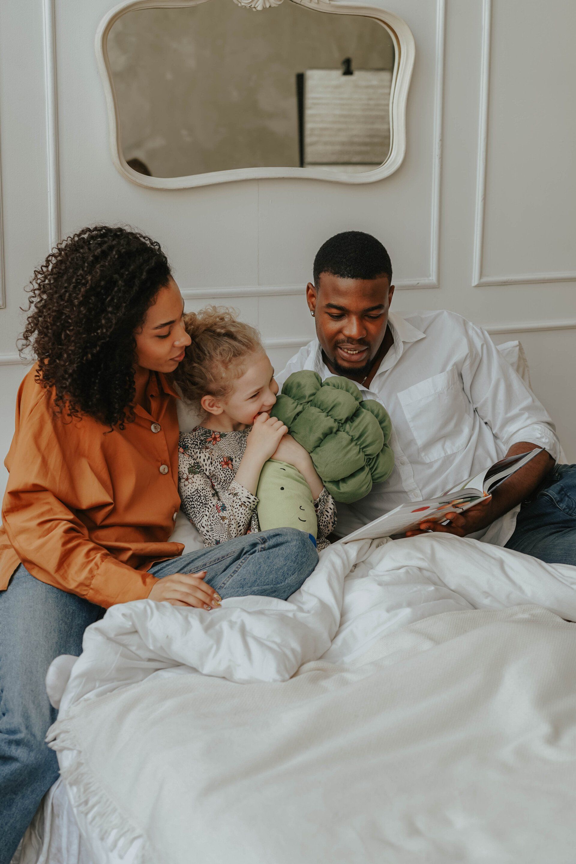 Family sitting on a bed, looking at a book together in a bright bedroom.