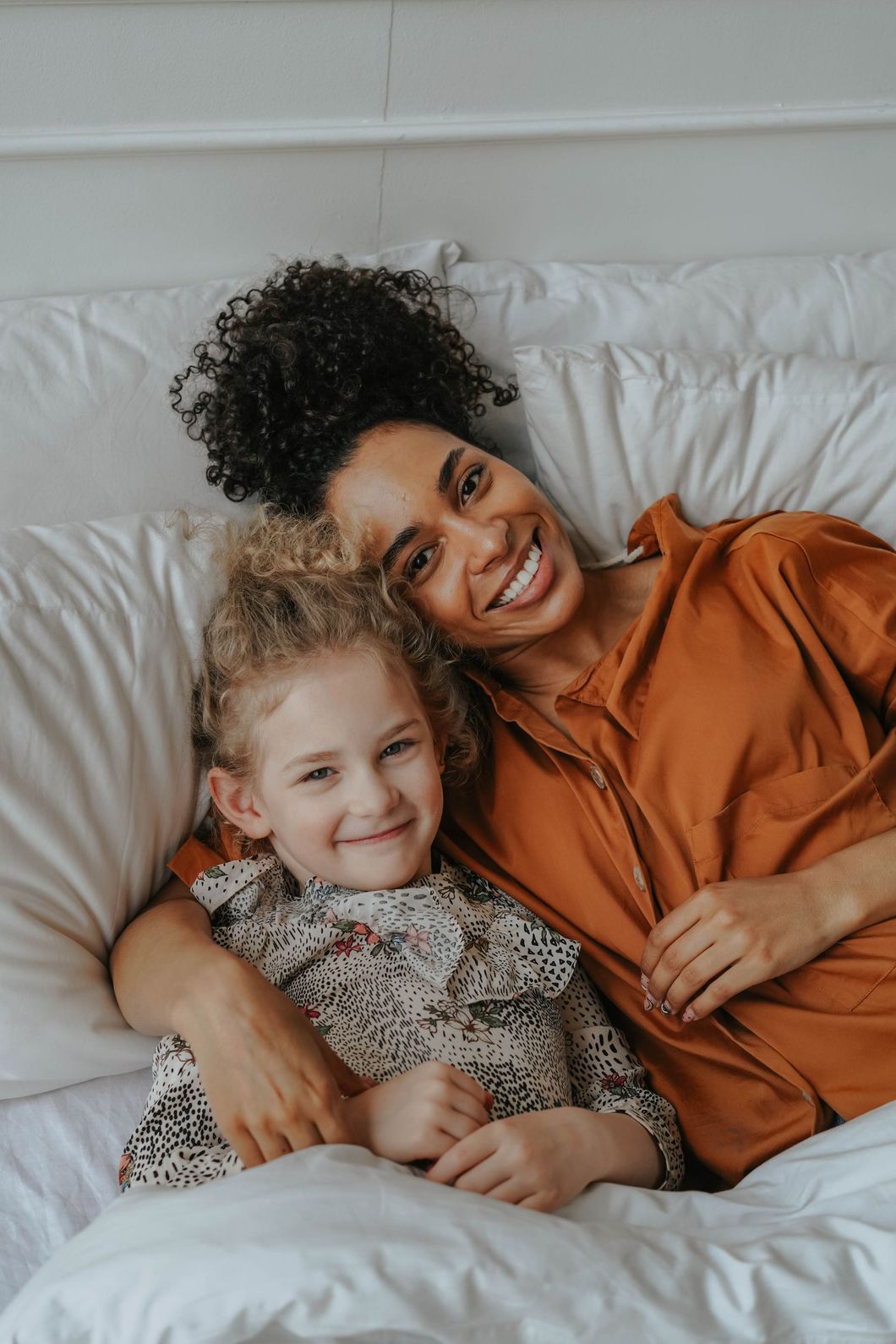 Woman with dark curly hair smiles at the camera, embracing a young girl with blonde curls, both lying on a white bed.