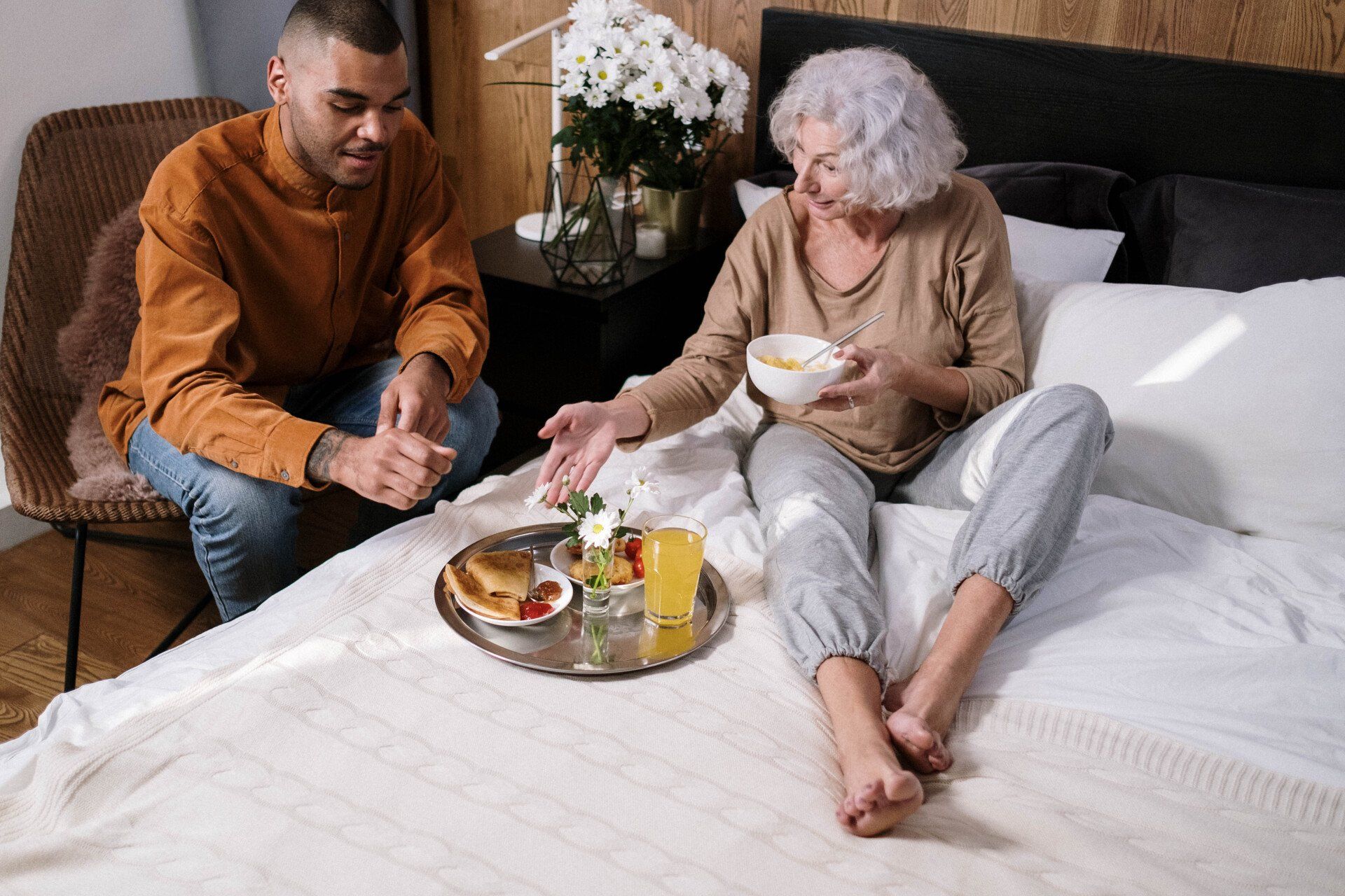 Man and person with gray hair on a bed, eating breakfast. Tray of food between them.