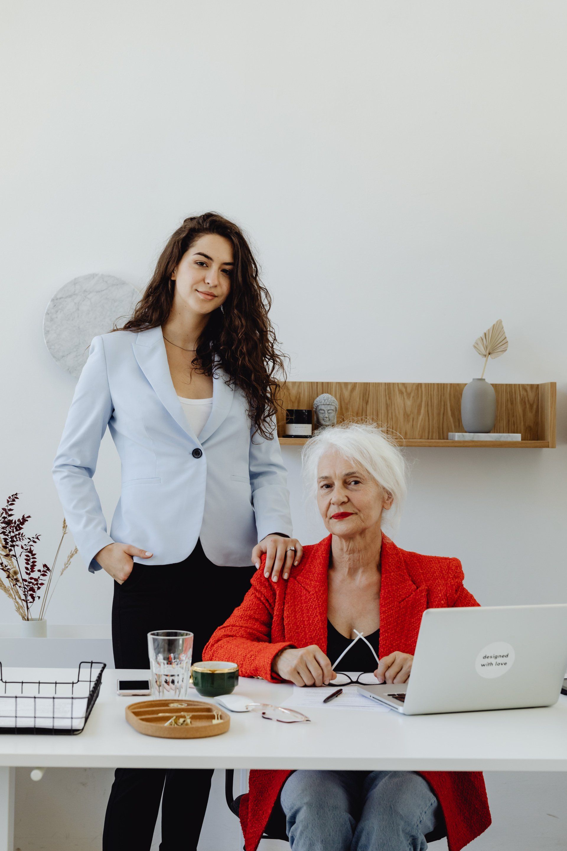 Een professional staat achter een zittende collega aan een wit bureau met een laptop; beiden kijken naar de camera.
