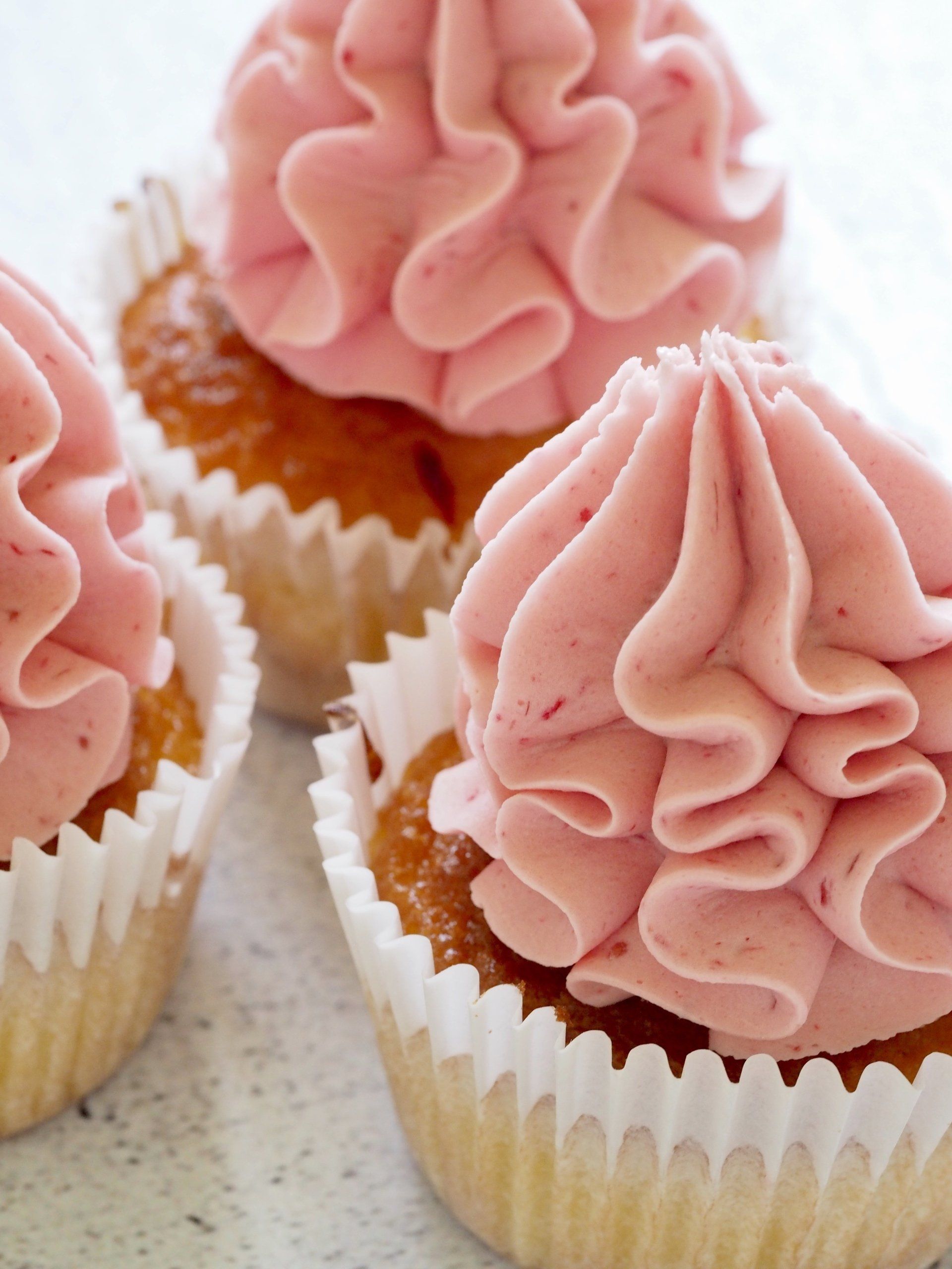 Three cupcakes in white paper liners, topped with tall, ruffled swirls of pale pink strawberry frosting.
