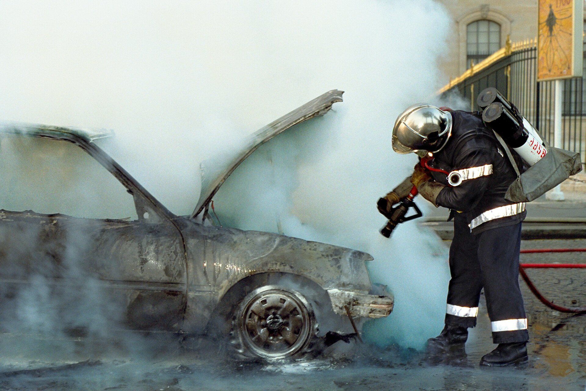 Firefighter spraying water on a car engulfed in smoke and flames outdoors