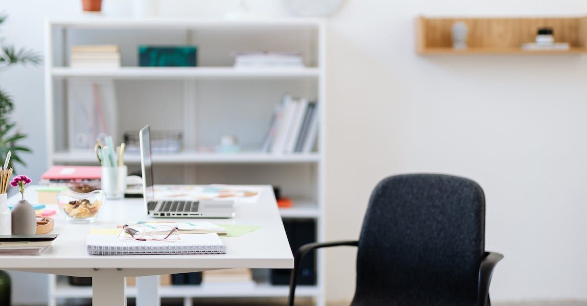 A fabric chair is at a white desk. On the desk are notebooks, stationary, and the laptops. Behind the desk is a white bookcase with a few books.