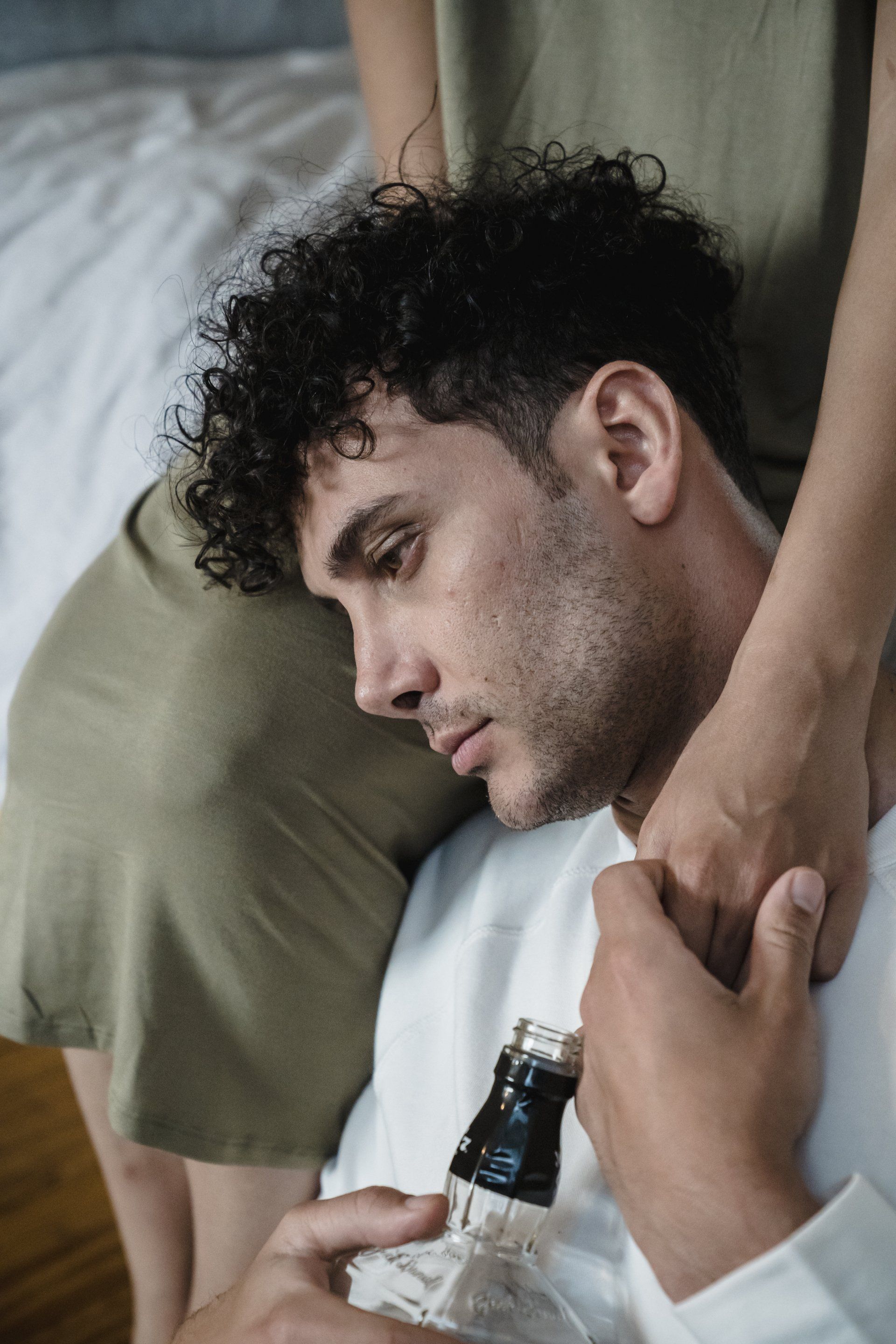 A man with curly hair rests his head on his hand, holding a liquor bottle. A person's arm rests on his shoulder, with a worried expression.
