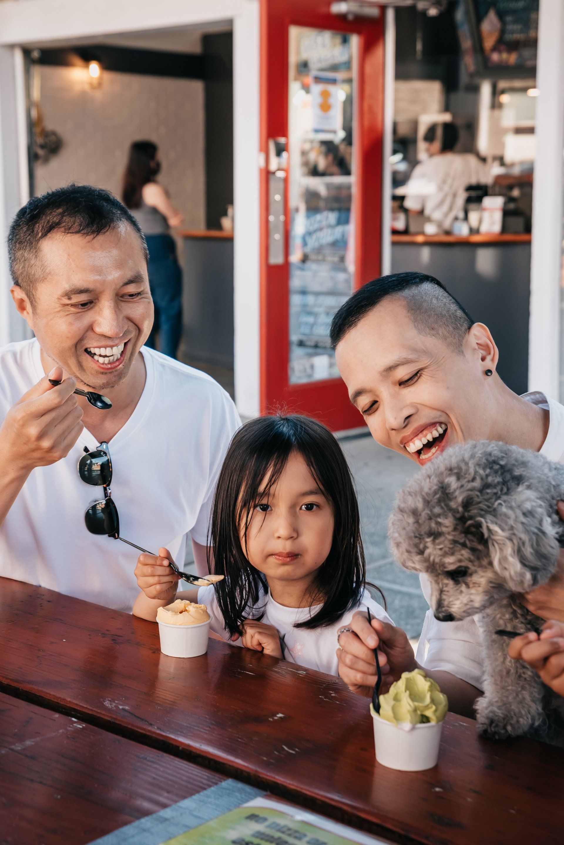 Two people and a small dog eating ice cream at a wooden table in front of a red-framed doorway.