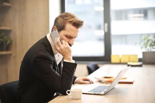 A man in a suit is talking on a cell phone while using a laptop computer.