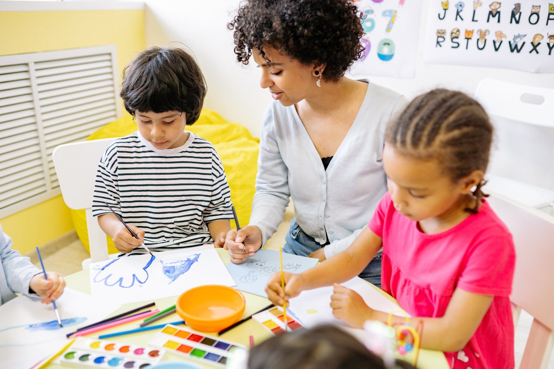 Teacher helps diverse children paint at a table in a bright classroom; colorful art supplies visible.