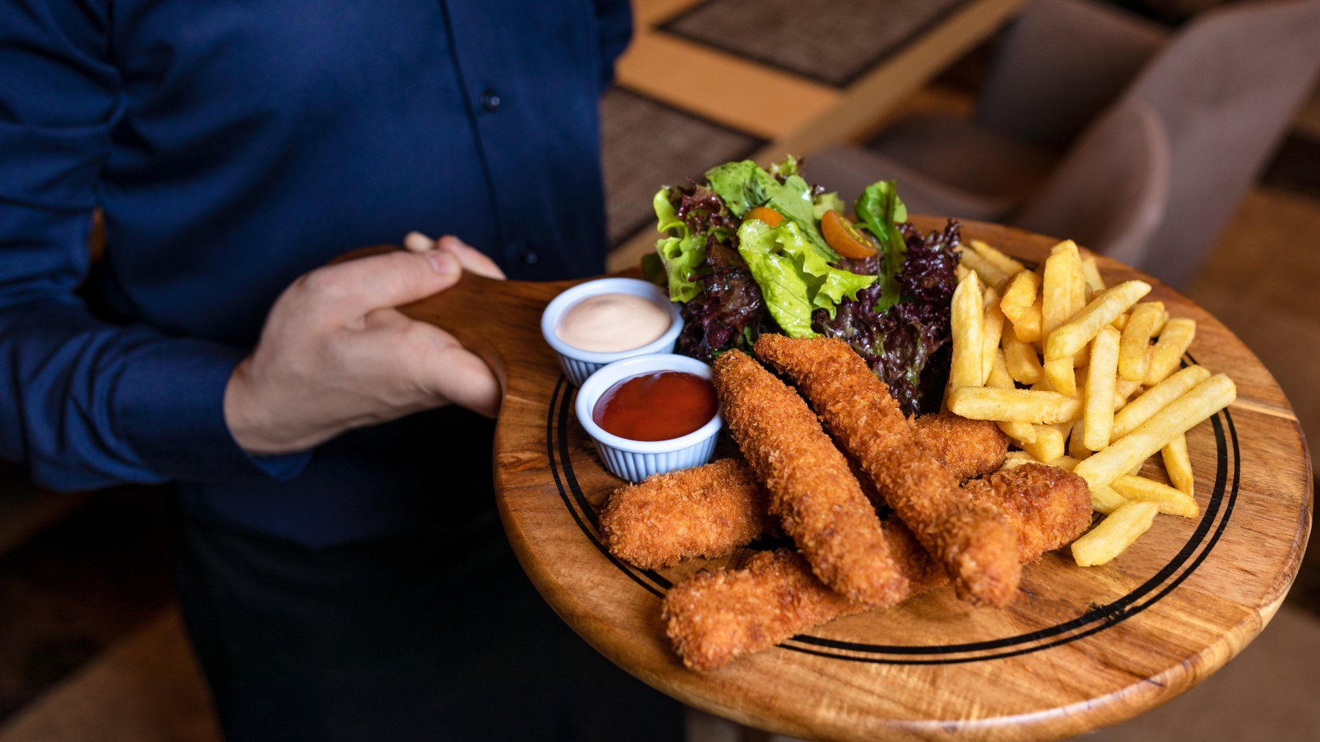 Person holding a wooden platter of fried appetizers, fries, broccoli, and dipping sauces at a restaurant table