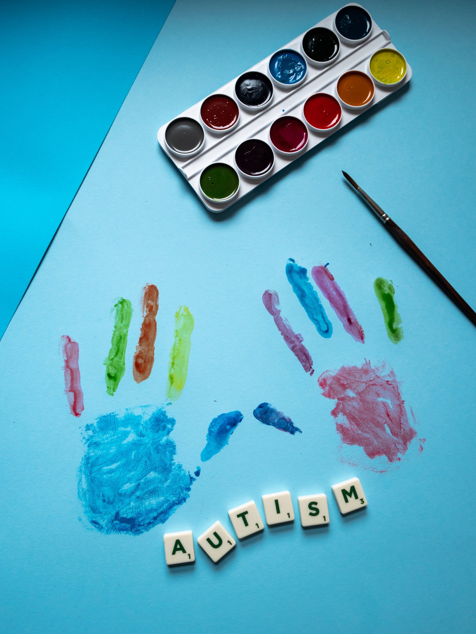 Aquarellpalette, Pinsel und bunte Handabdrücke neben Buchstabenplättchen, die „AUTISM“ ergeben