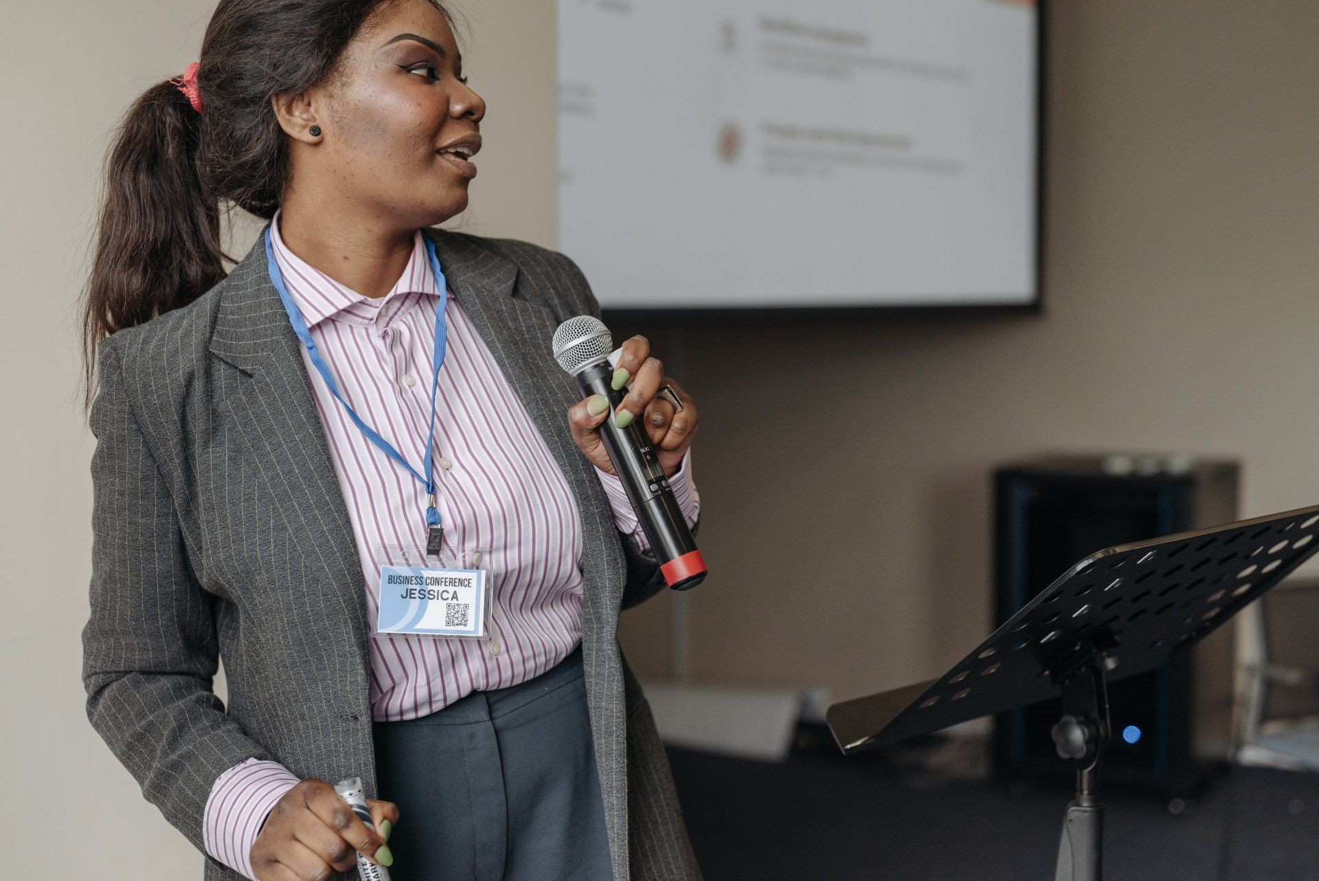Speaker holding a microphone at a podium in a conference room, wearing a gray blazer and striped shirt