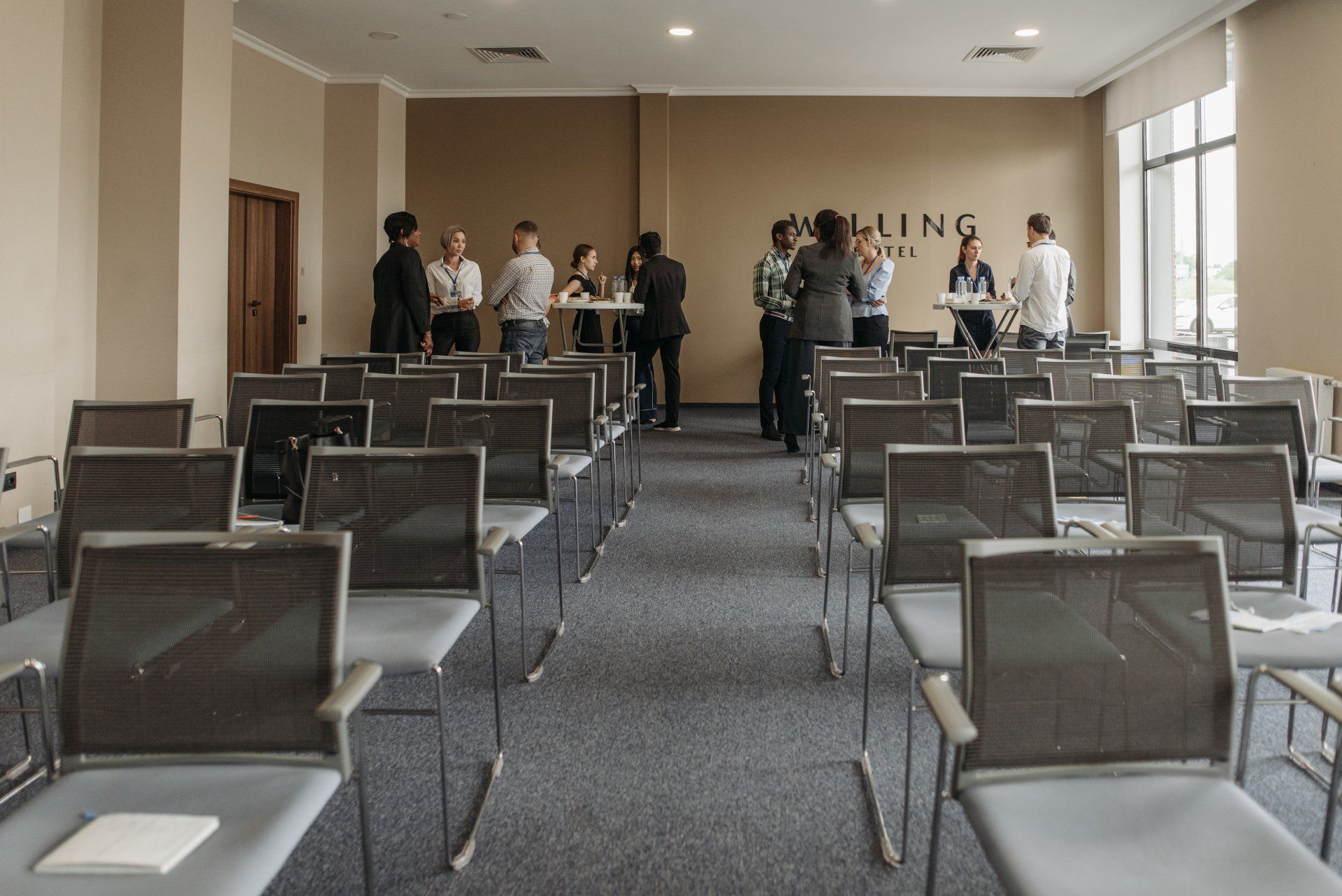 a group of people are standing around tables and chairs in a conference room .