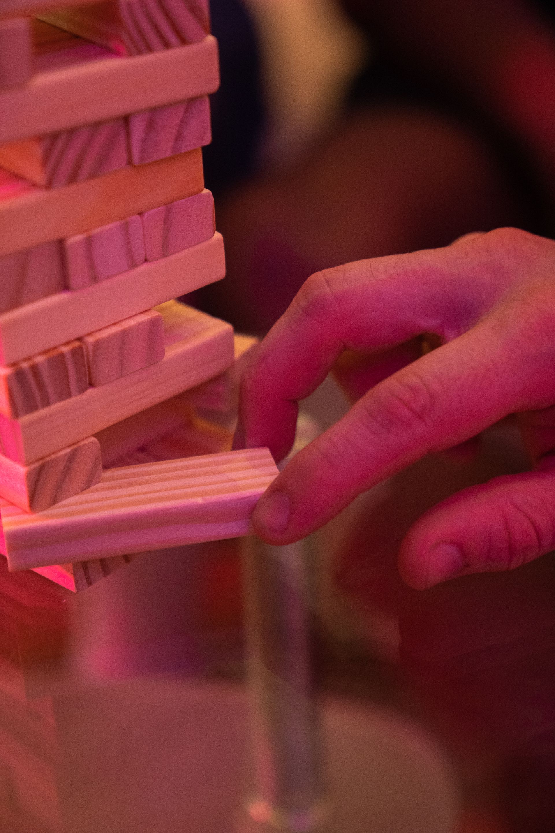 A person's hand carefully removing a wooden block from a Jenga tower, lit with pink light.