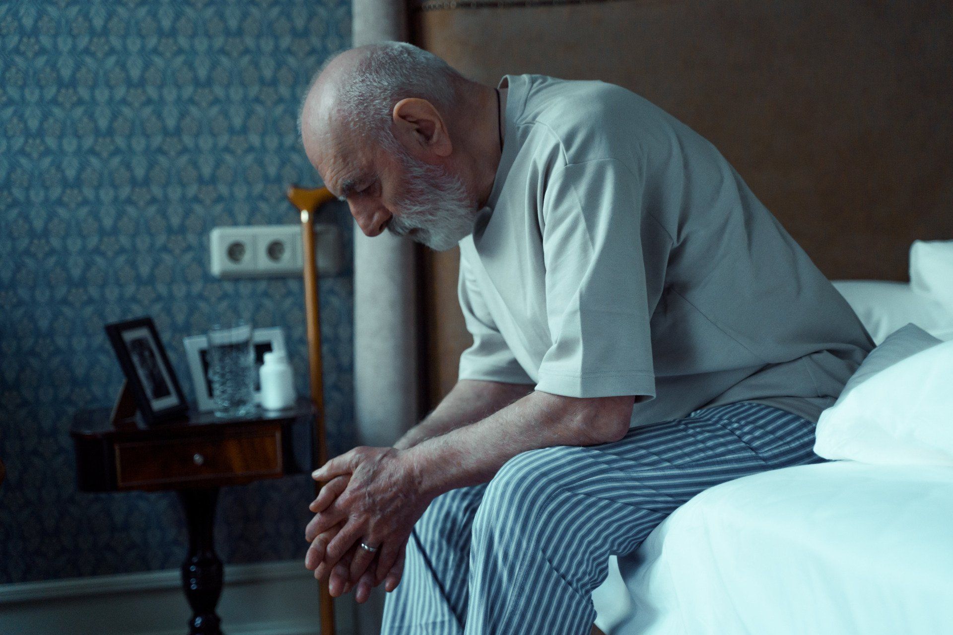 an older man in pajamas looking contemplative on the edge of his bed. A nightstand with a photo, glass, medication and cane are in the background