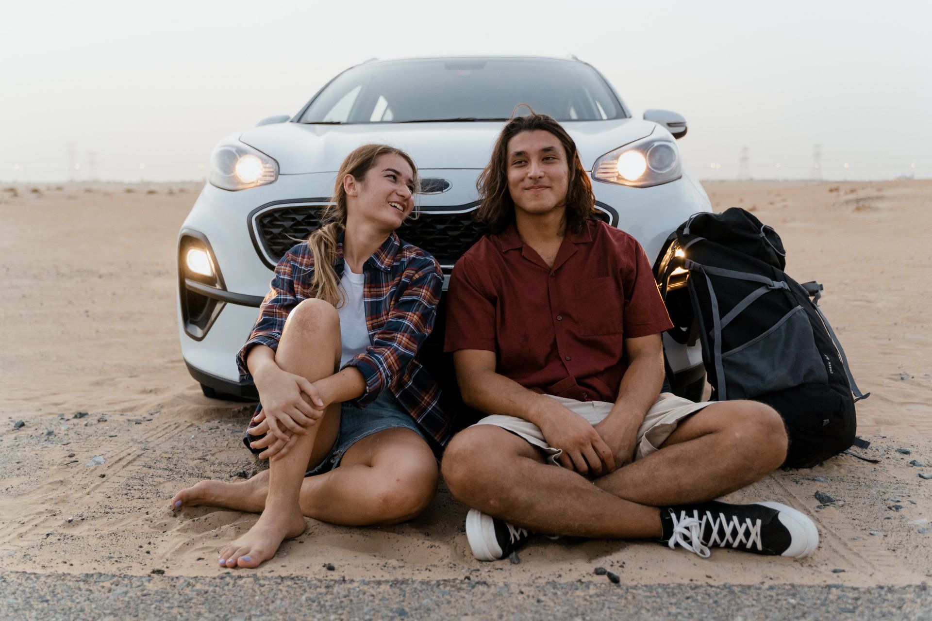 Two people sitting cross-legged in front of a car in a sandy desert, smiling at each other.