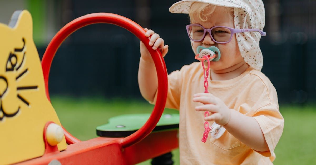 A child wearing a hat and glasses holds a pacifier attached to a pink chain while standing by a yellow playground toy.