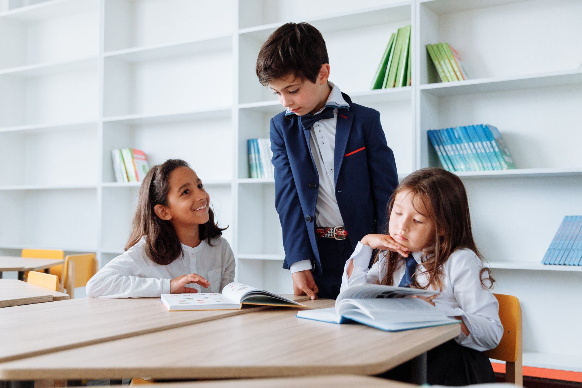 A boy in a suit is standing next to two girls in a classroom.