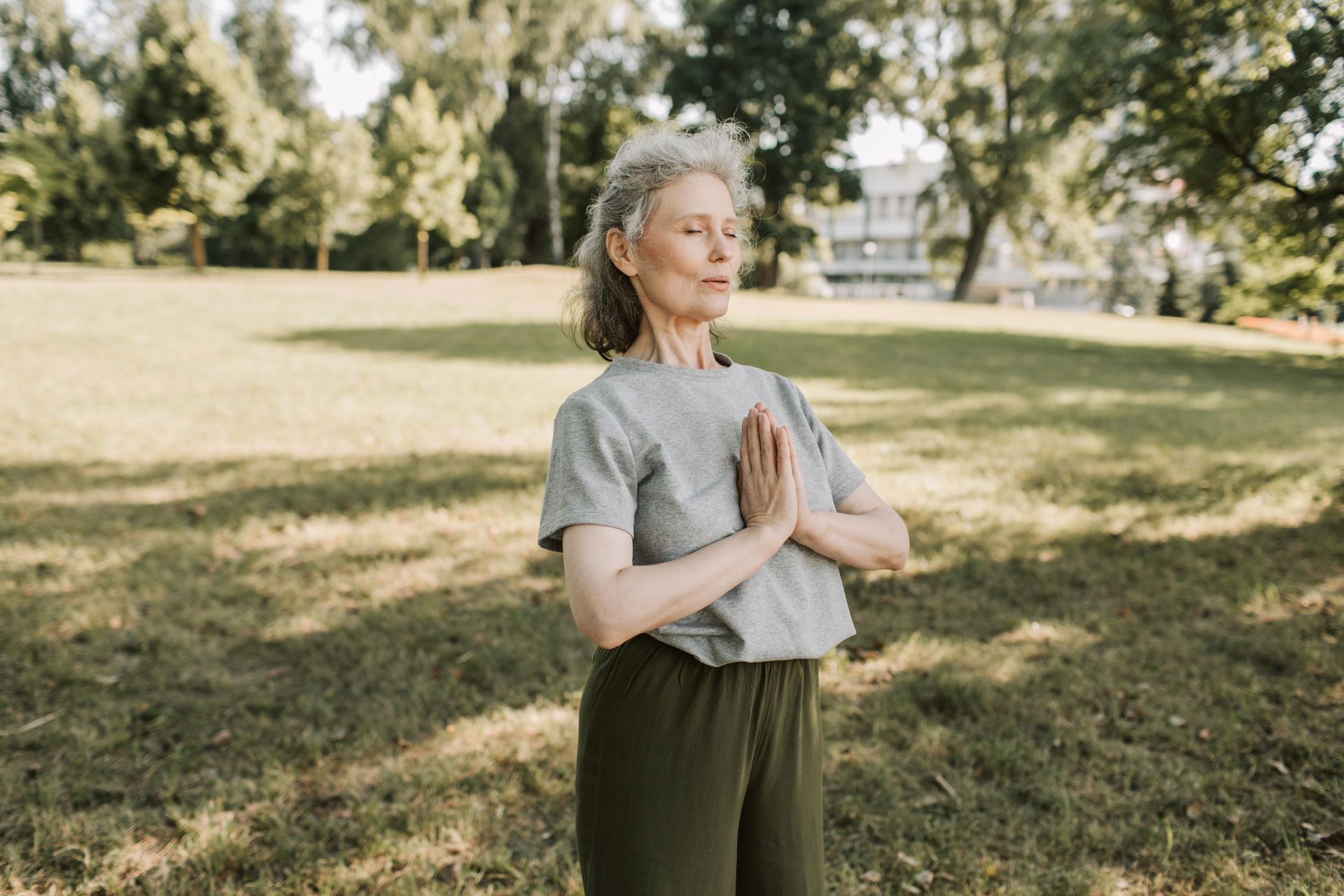 Elderly woman in a park with hands clasped, eyes closed, practicing yoga outdoors.