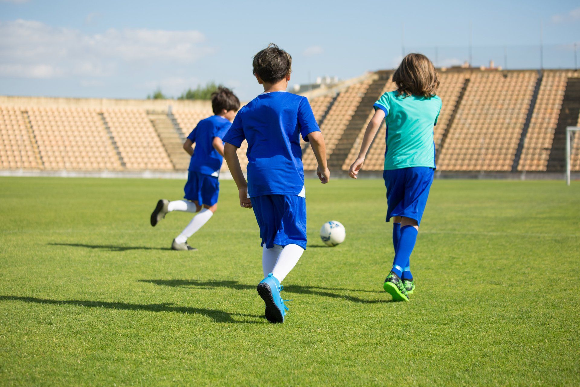 Boys playing soccer on a green field with a stadium in the background.